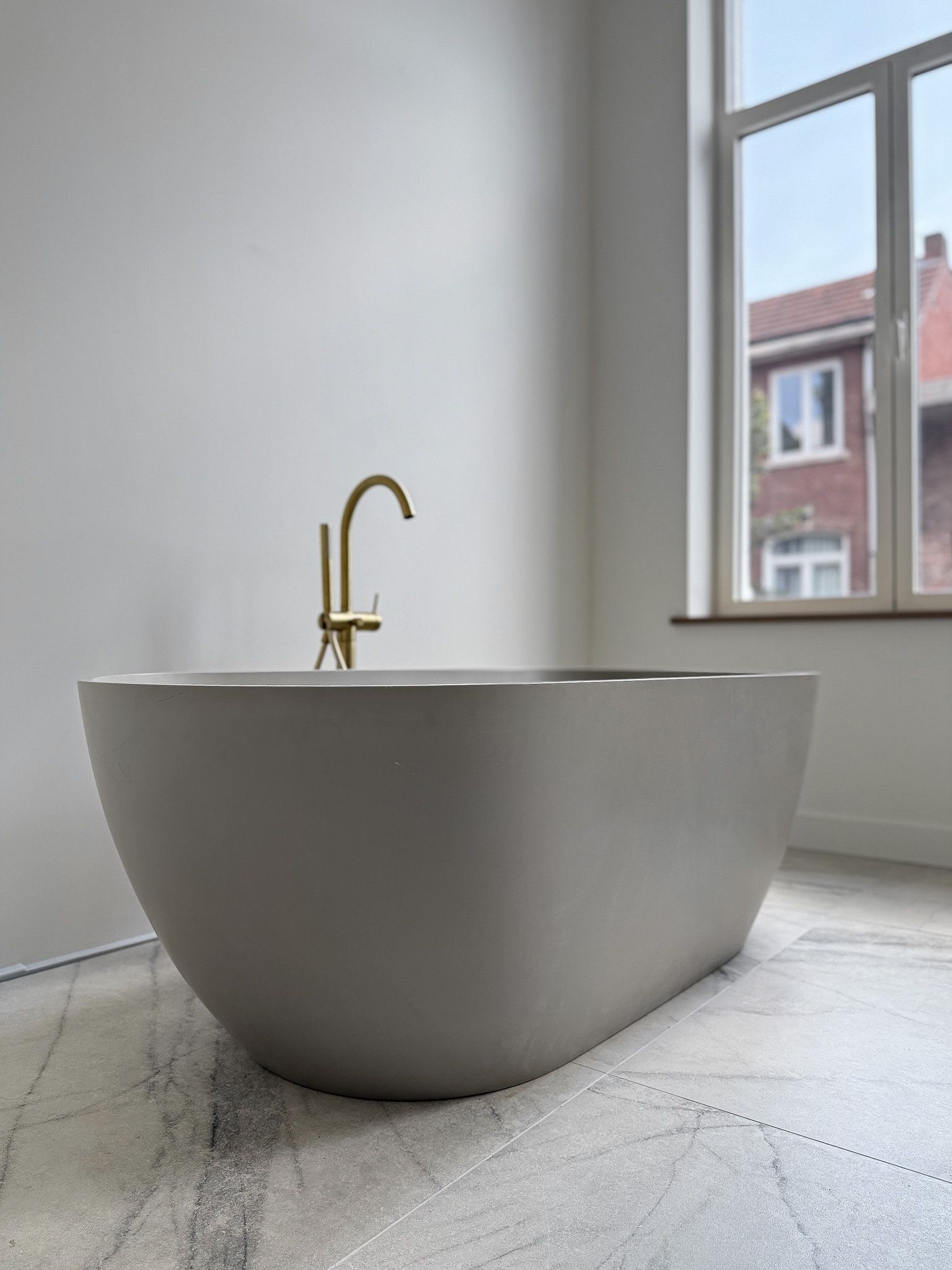 Vrijstaand grijs bad met gouden kraan in een moderne badkamer; een raam biedt uitzicht op een rood bakstenen gebouw.