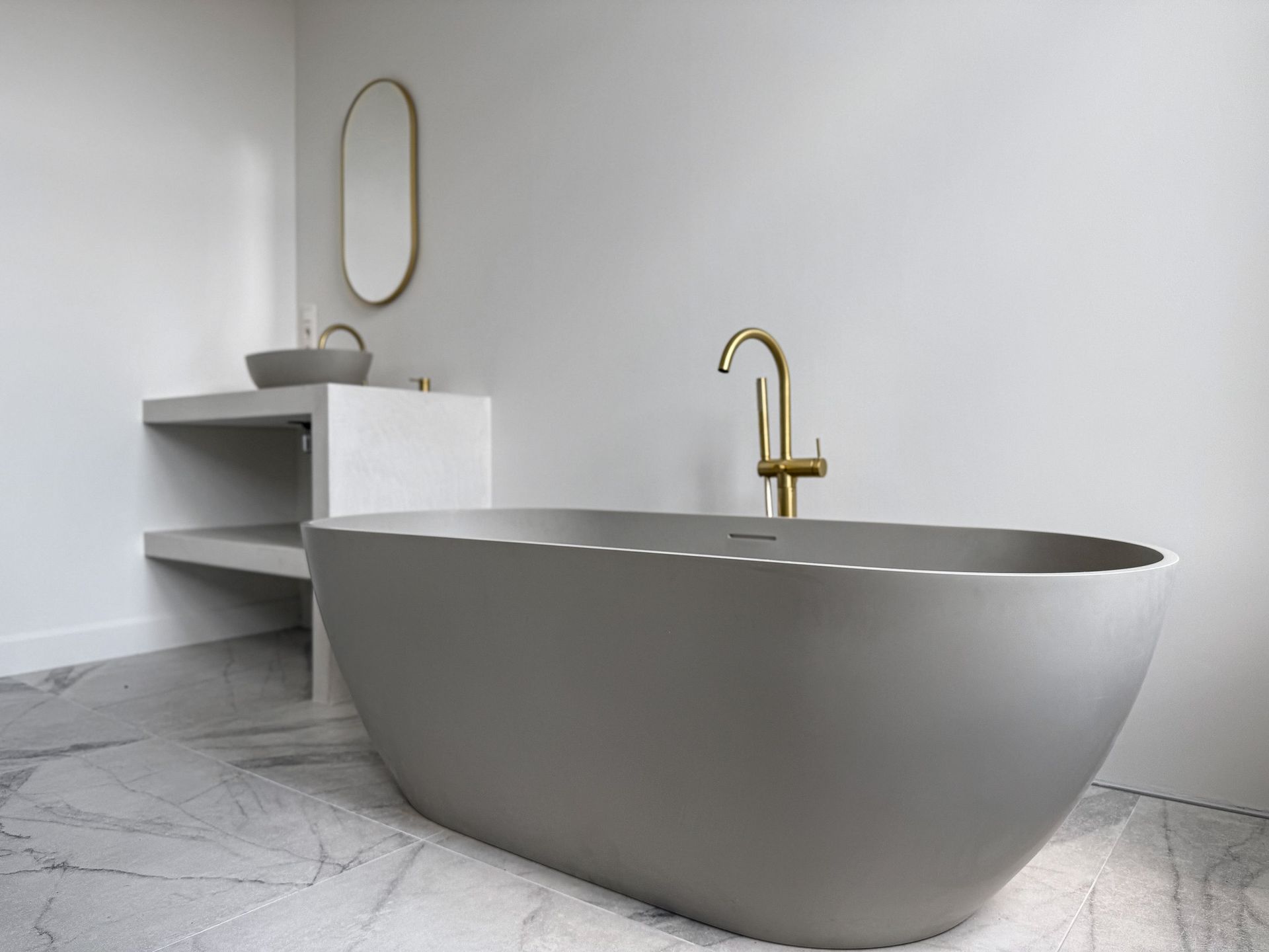 Moderne badkamer met een grijs ovaal bad, gouden kraan en wastafel op een marmeren vloer.