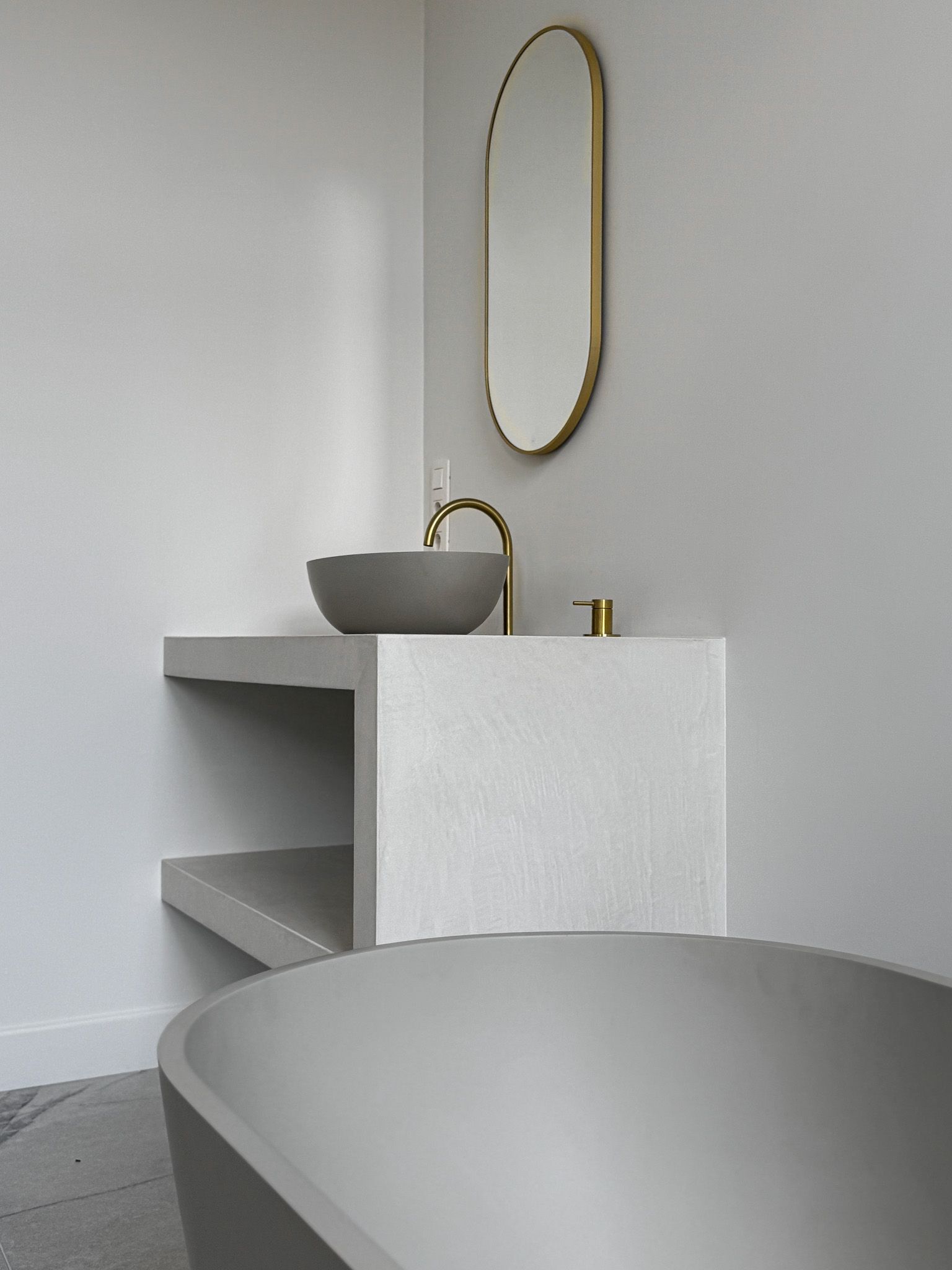 Moderne grijze badkamer met een betonnen wastafel, gouden spiegel en bad.
