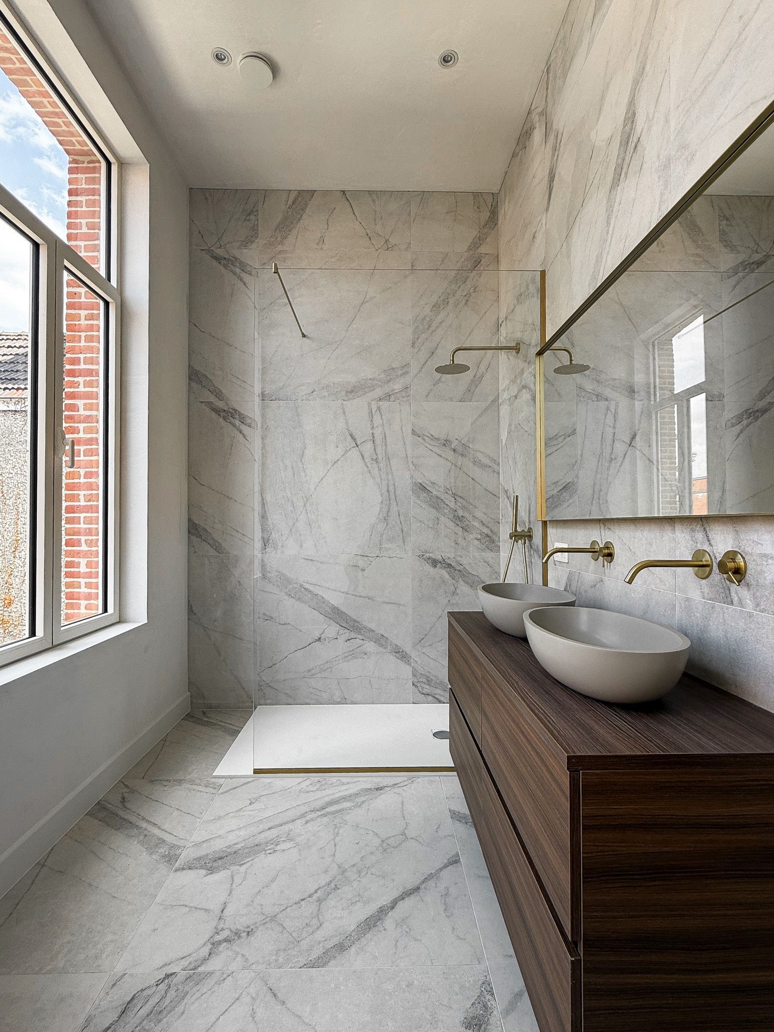 Moderne marmeren badkamer met een groot raam, houten wastafel en gouden armaturen.