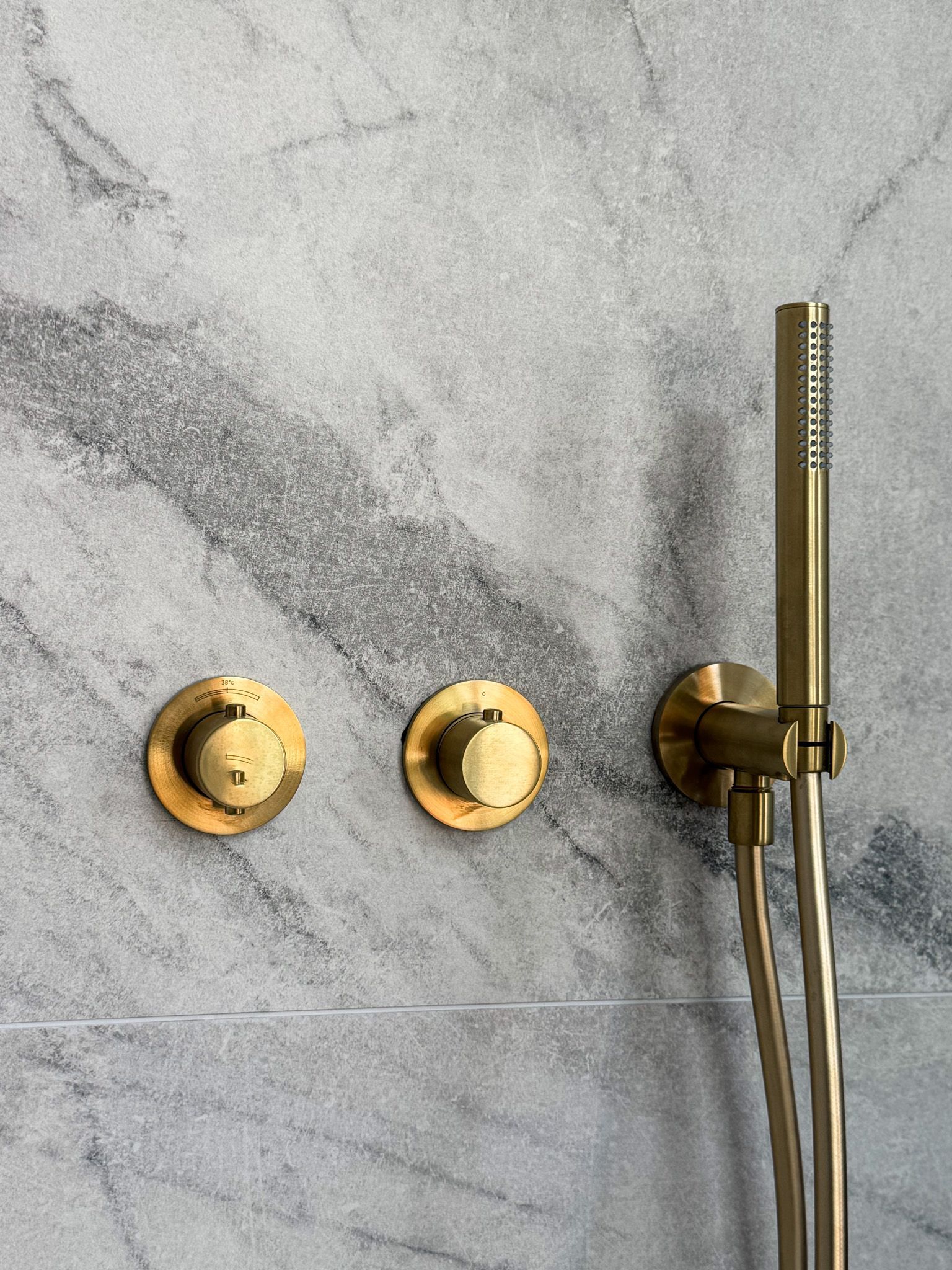 Gouden douchearmaturen op een grijze marmeren muur; twee ronde knoppen en een handdouche.