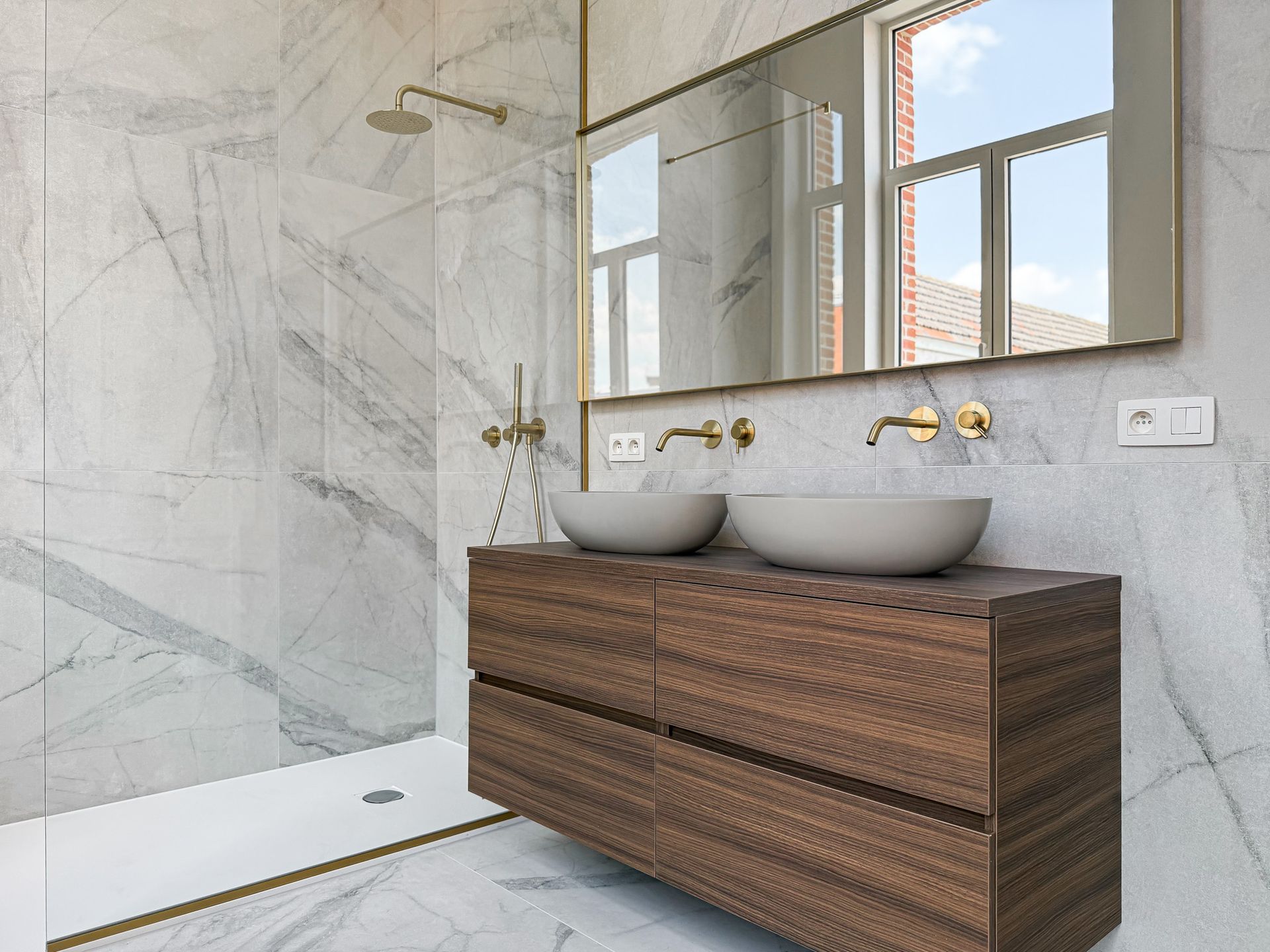 Moderne badkamer met marmeren muren, houten wastafelmeubel, twee opzetwastafels en gouden armaturen.