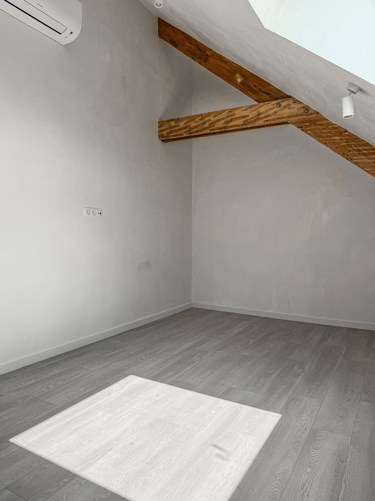 Lege kamer met grijze muren, zichtbare houten balken en grijze vloer. Op de vloer staat een zonnig vierkant.