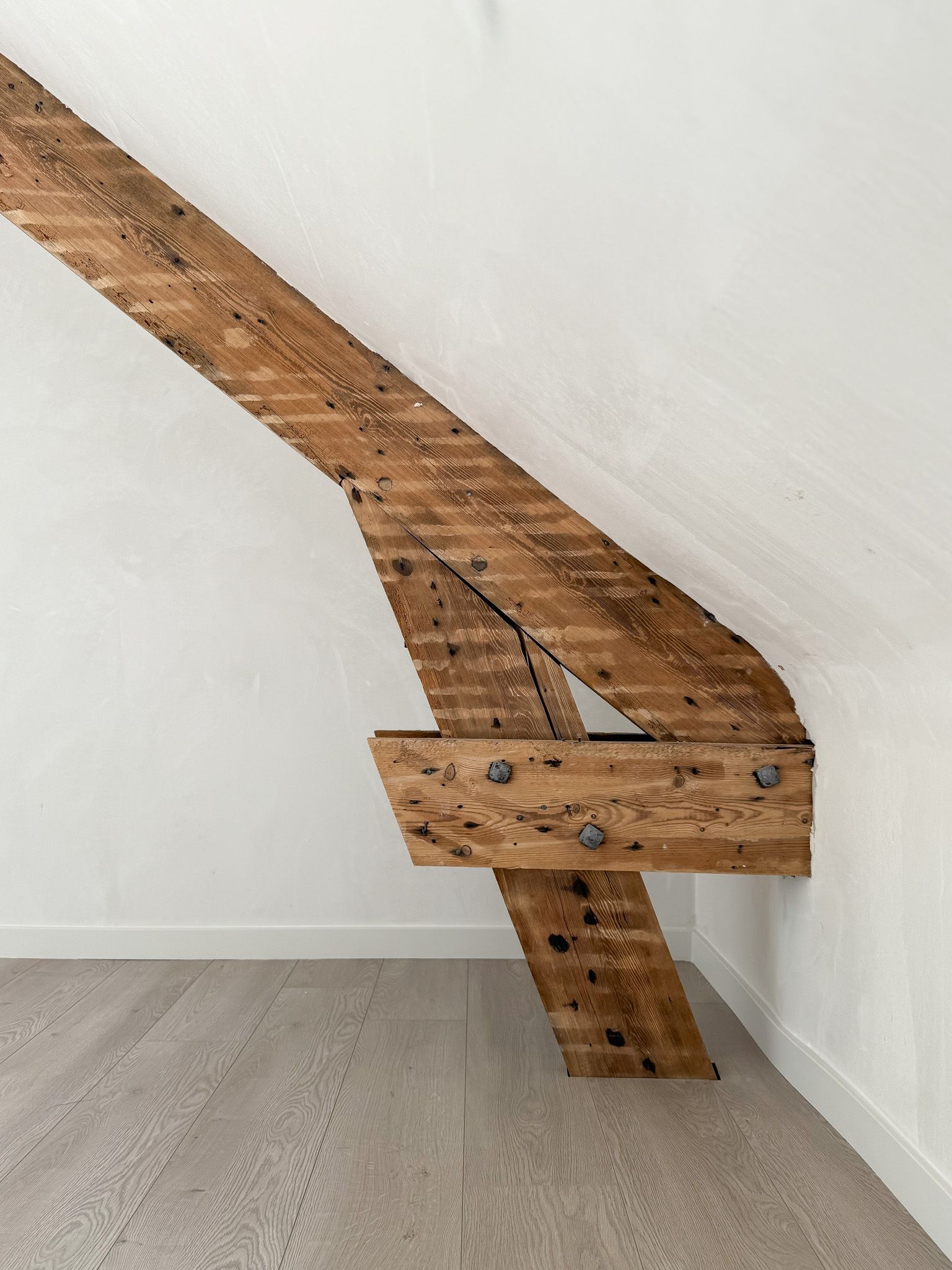 Houten steunbalk bevestigd aan een witte muur. Bruin hout en grijze vloer.