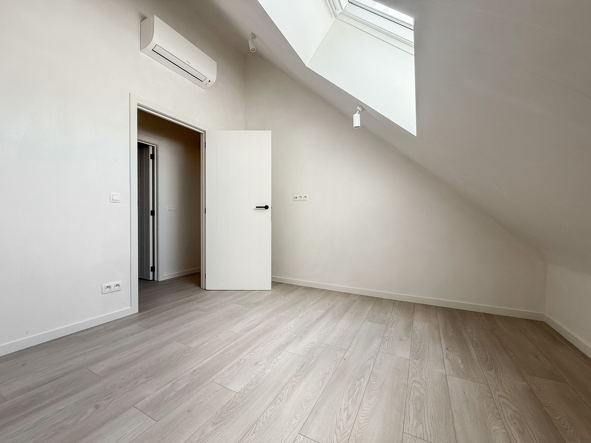 Lege zolderkamer met witte muren, schuin plafond, dakraam en lichte houten vloeren.