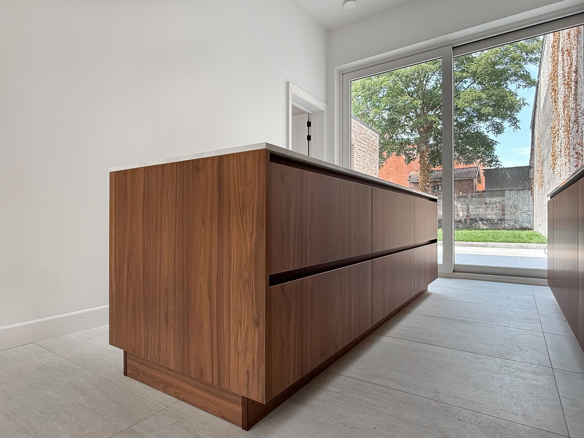 Keukeneiland van hout met lades, een wit aanrechtblad en een raam met uitzicht op een bakstenen muur.