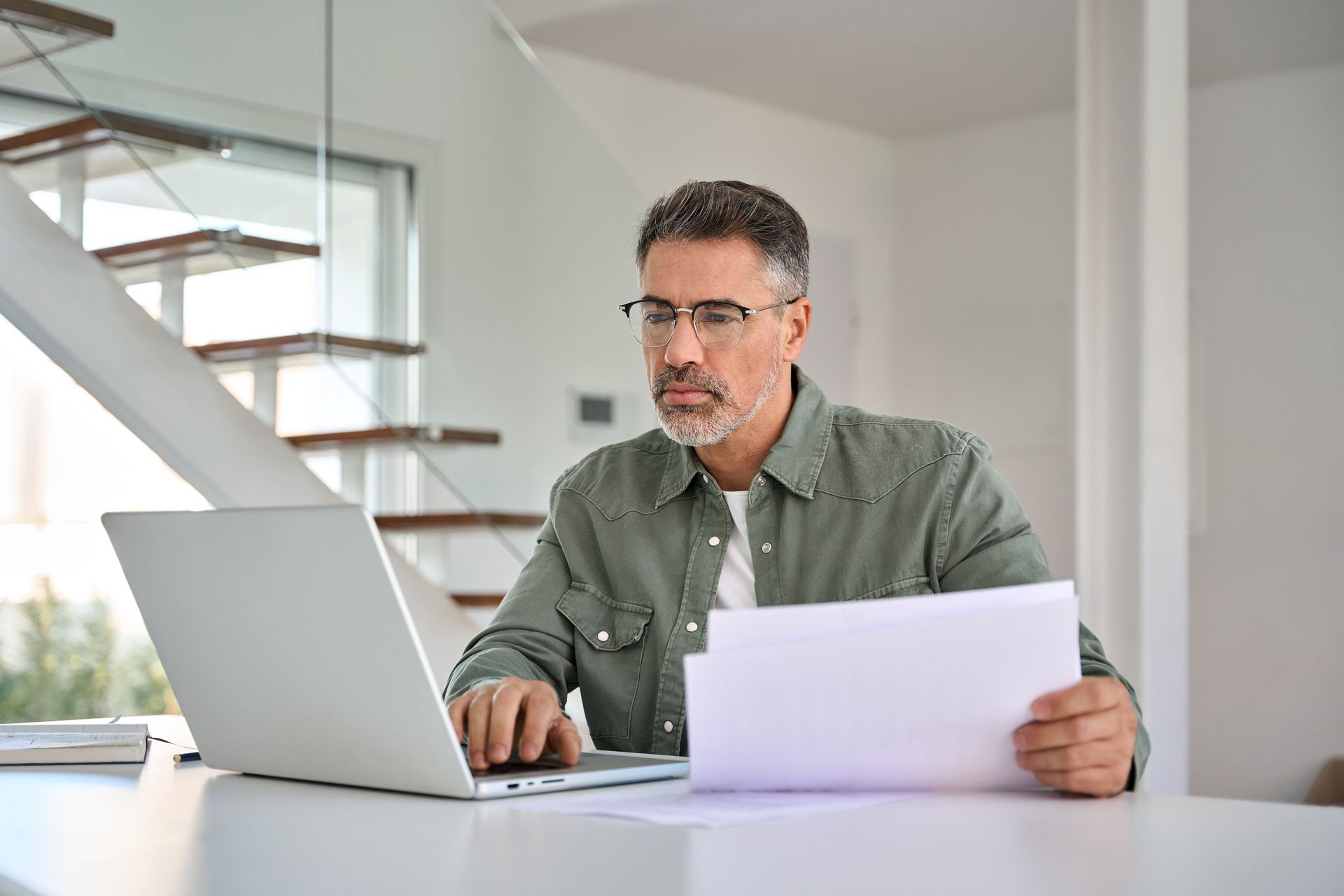 Man met bril kijkt op een laptop en houdt papieren vast, binnenshuis bij een trap.
