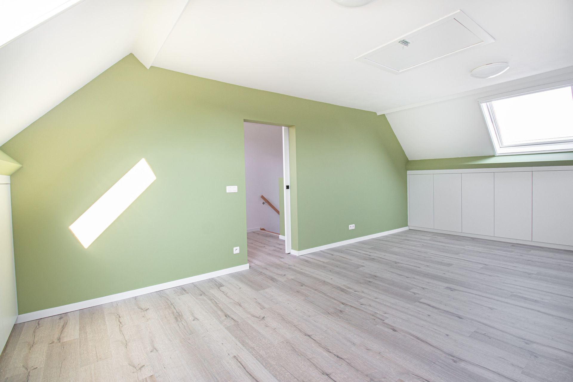Zolderkamer met groene muren, grijze vloer, witte kasten en een deur naar de trap.