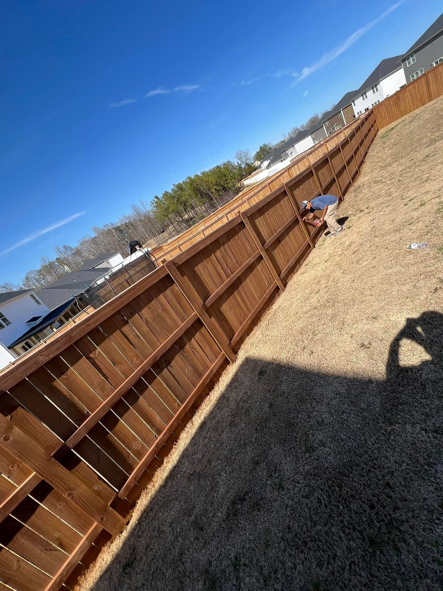 A person stands by a long, wooden privacy fence in a backyard under a clear blue sky.