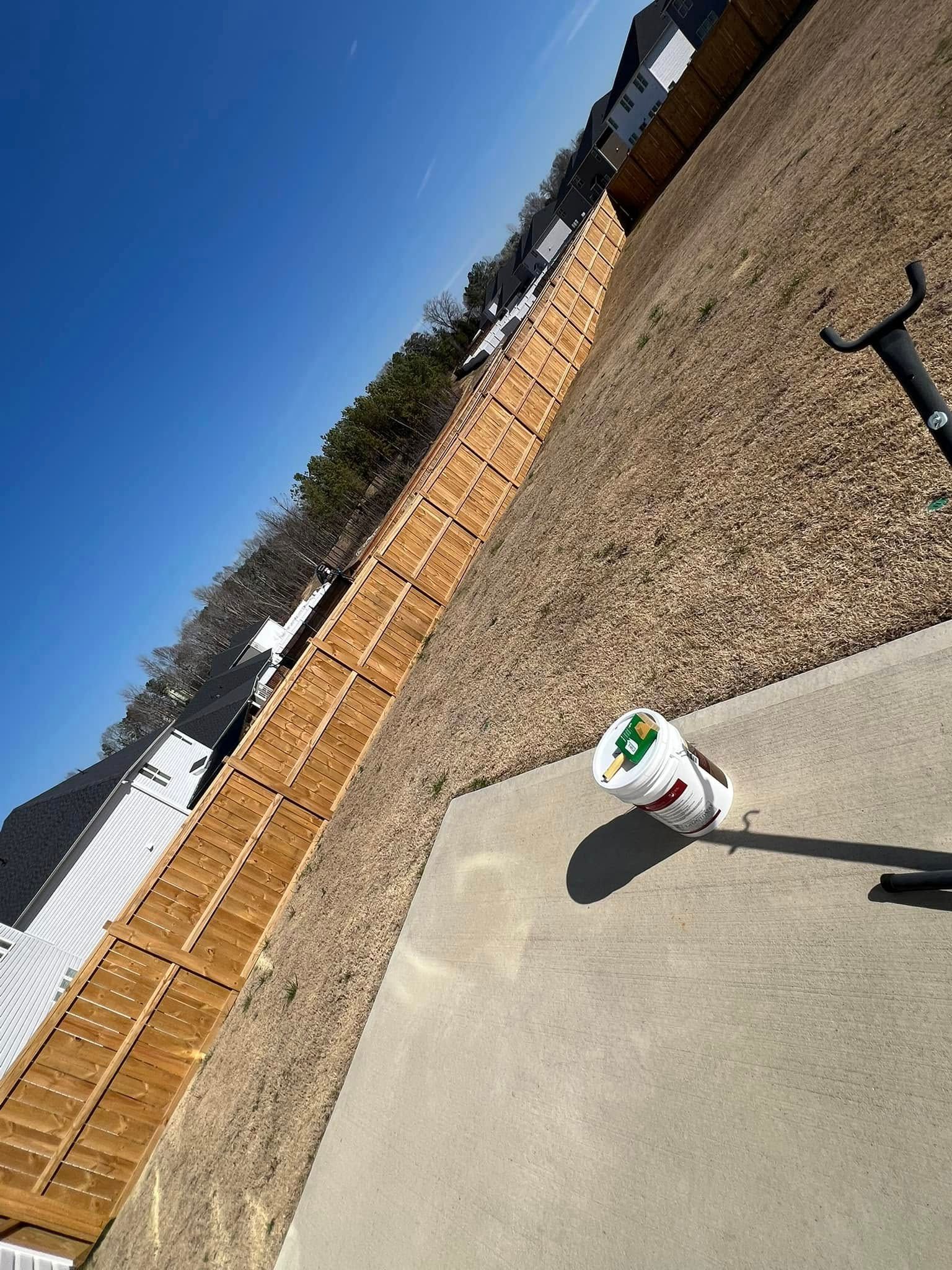 A bright backyard with a wood fence, a patch of dry grass, and a white bucket resting on a concrete patio.