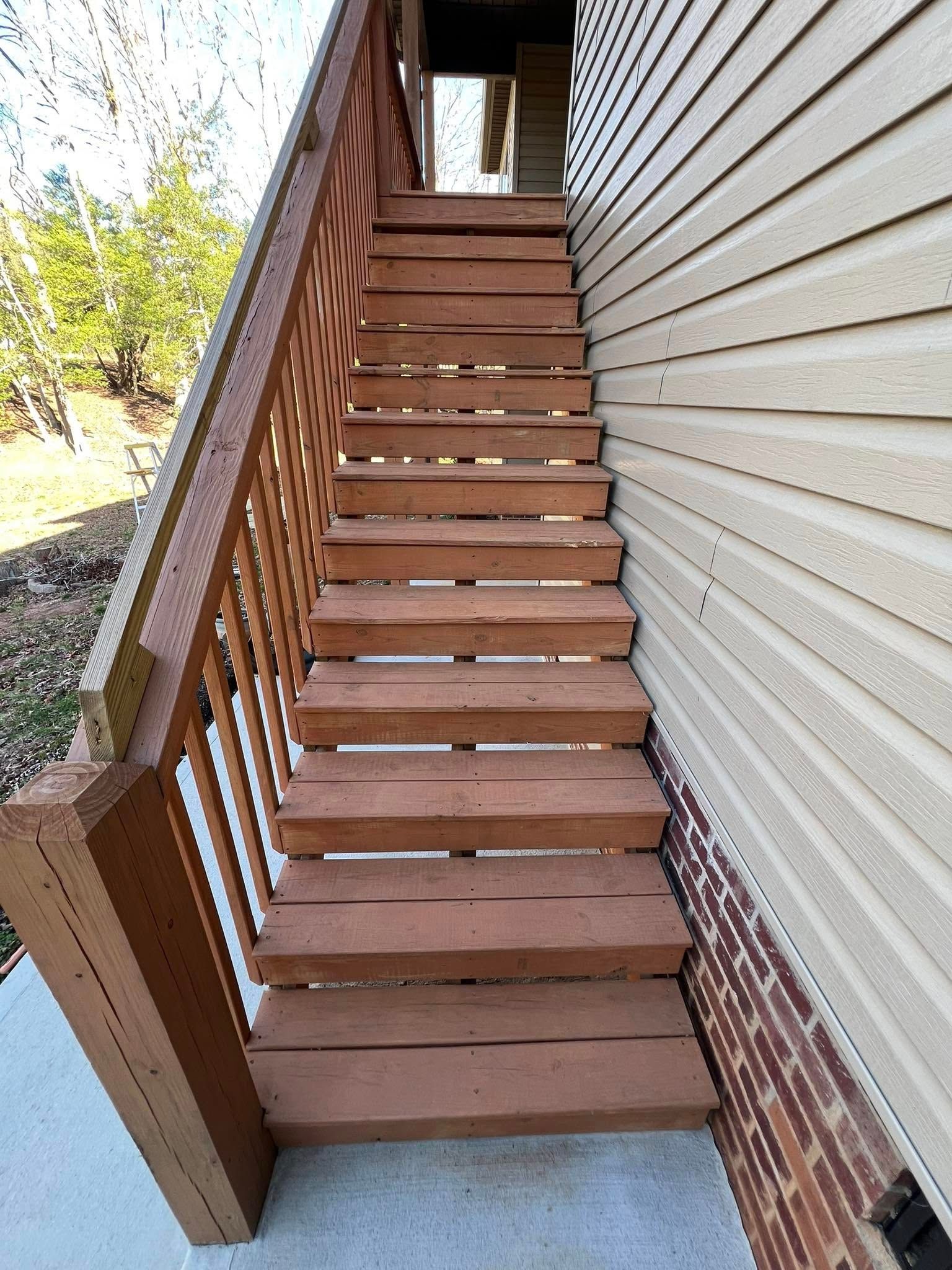 A wooden staircase with a railing leads up the side of a building with tan siding and a brick base.