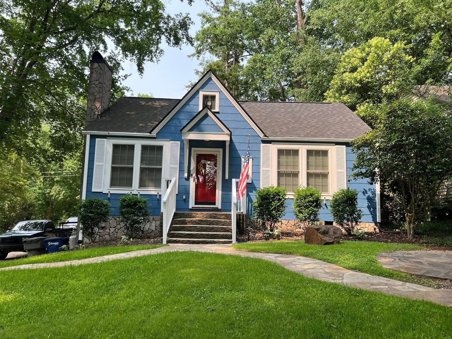 A blue, one-story cottage with a stone foundation, a red front door, and a grassy front yard surrounded by trees.