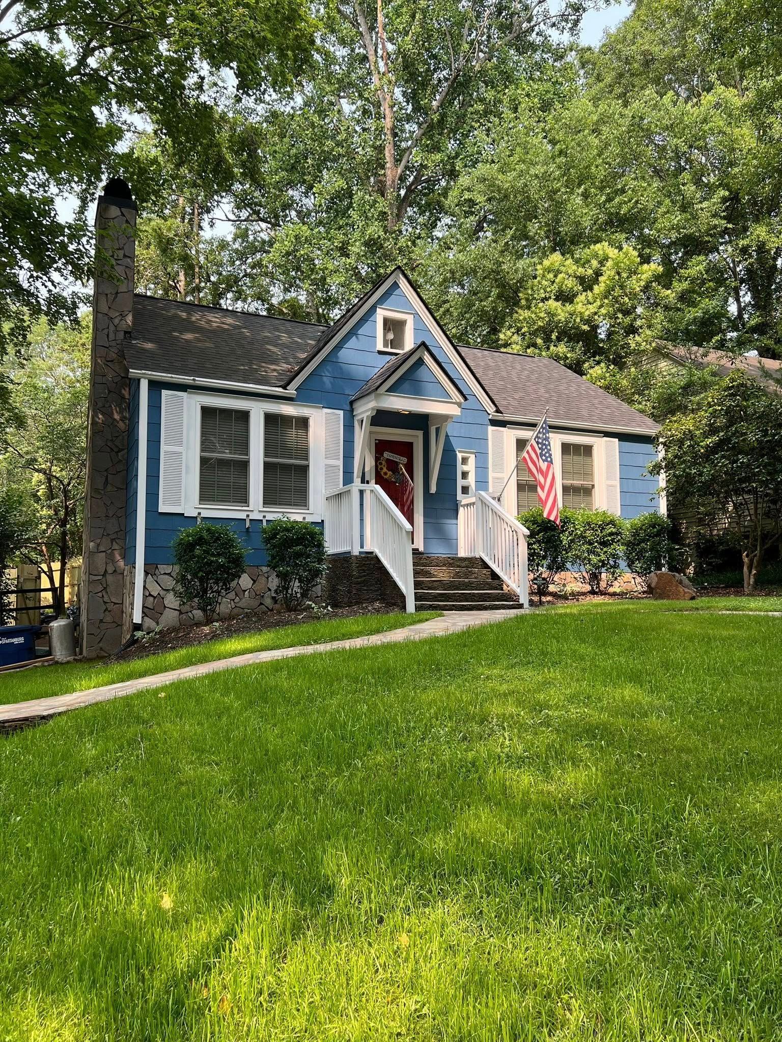A charming blue house with white trim and a stone foundation, set in a lush green yard surrounded by mature trees.
