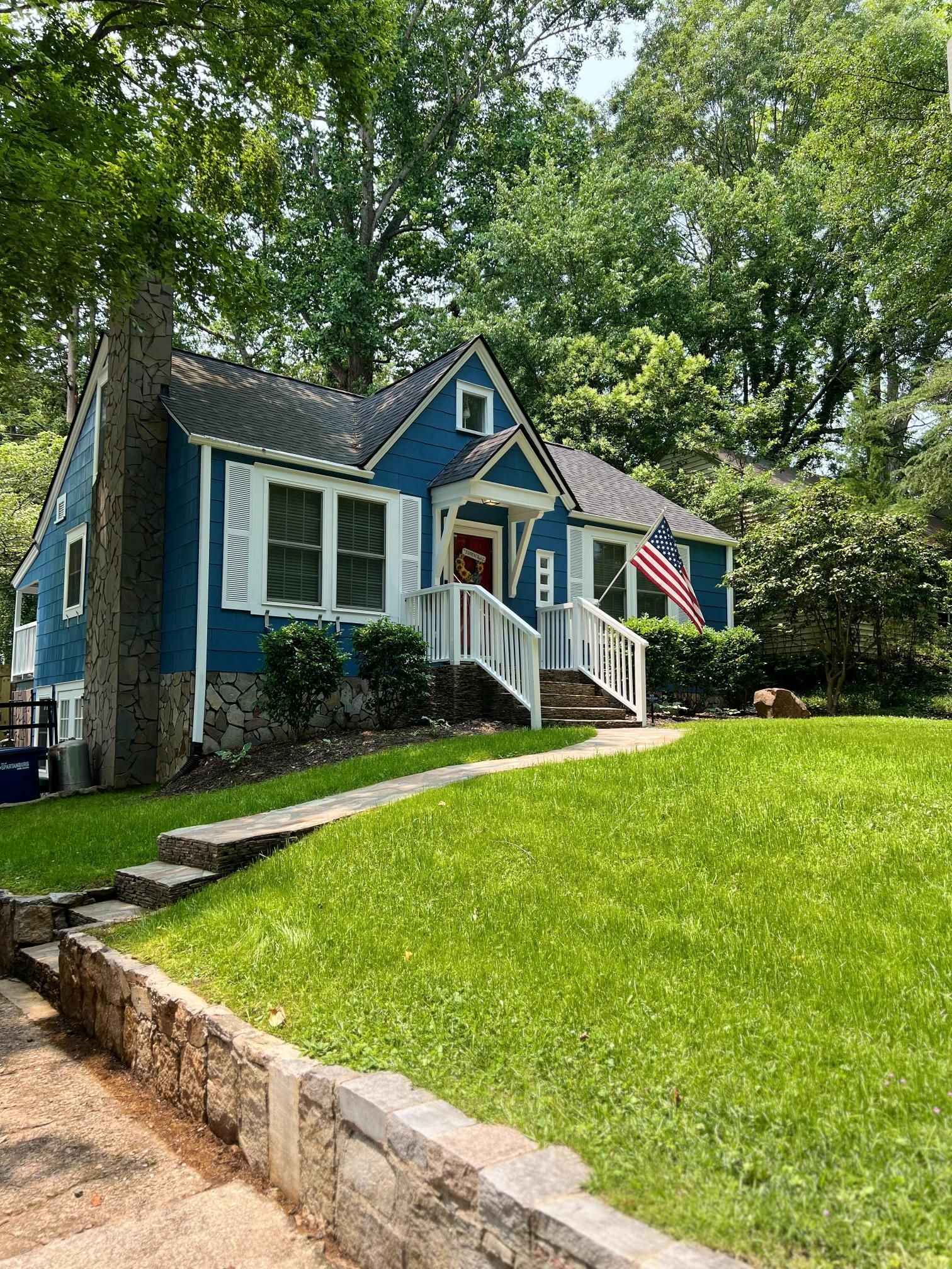 A blue house with white trim and a stone foundation sits on a grassy hill, accessed by a stone staircase.