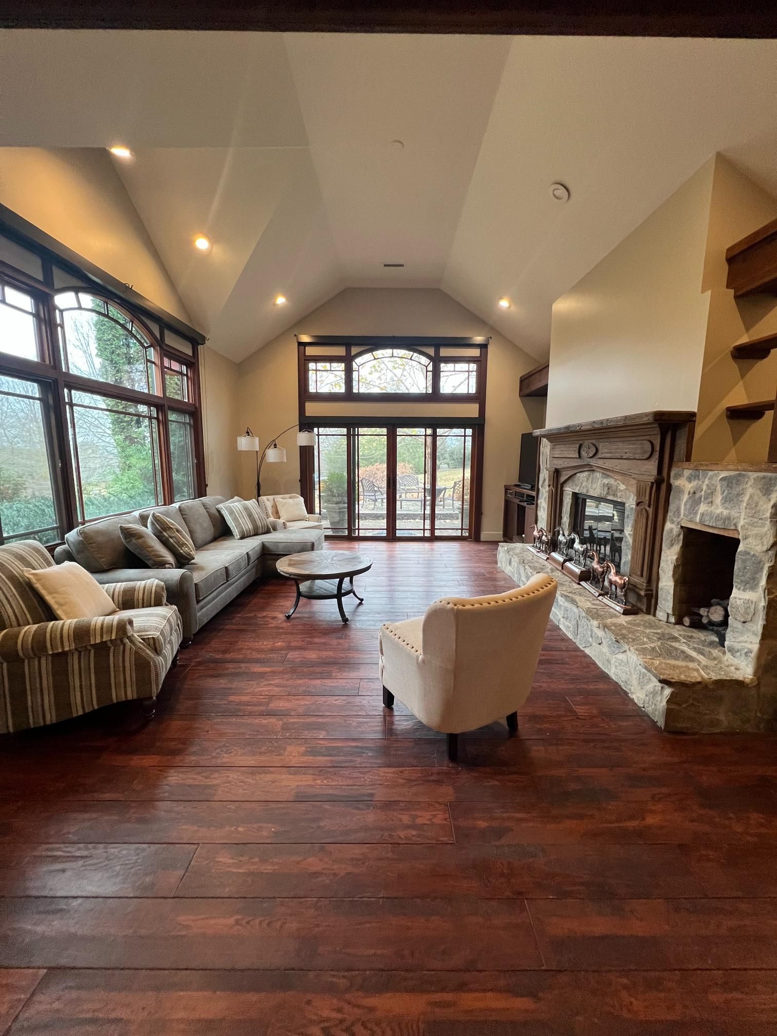 A spacious living room with dark wood floors, a stone fireplace, large windows, and comfortable seating under a high ceiling.