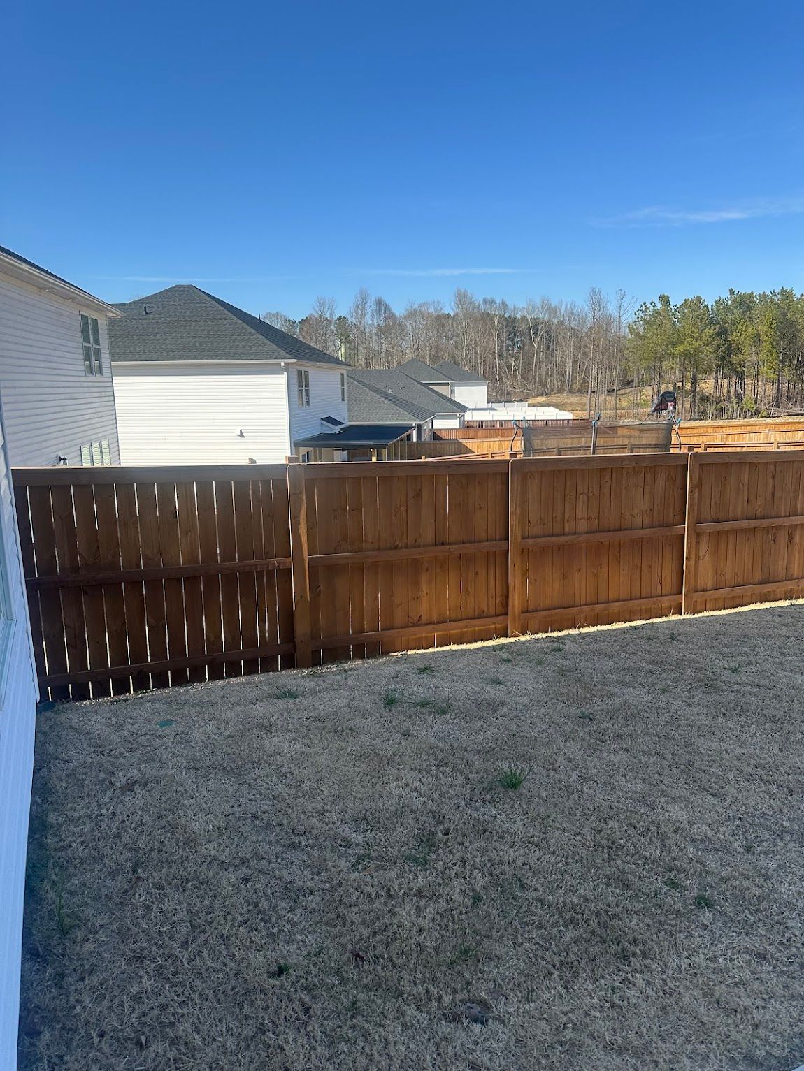 A backyard with a wooden privacy fence, sparse grass, and a view of neighboring houses under a clear blue sky.