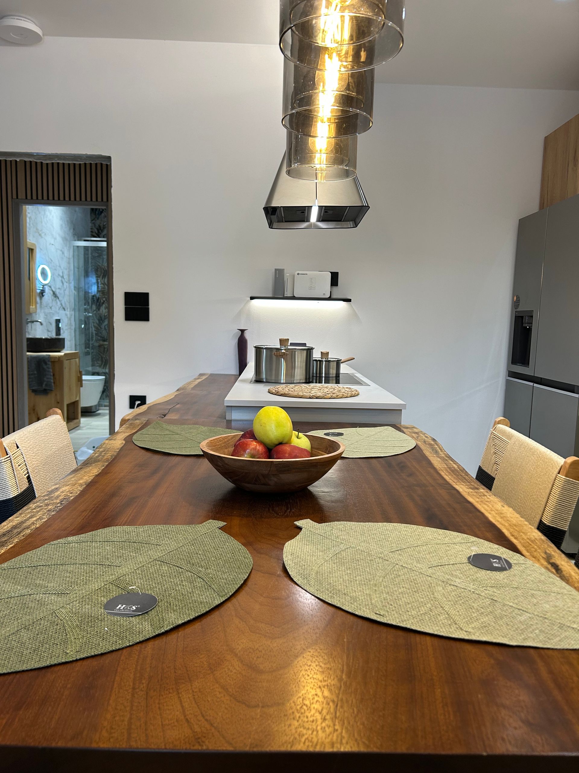 Dining table with placemats and fruit bowl in a modern kitchen with a range hood and stove in the background