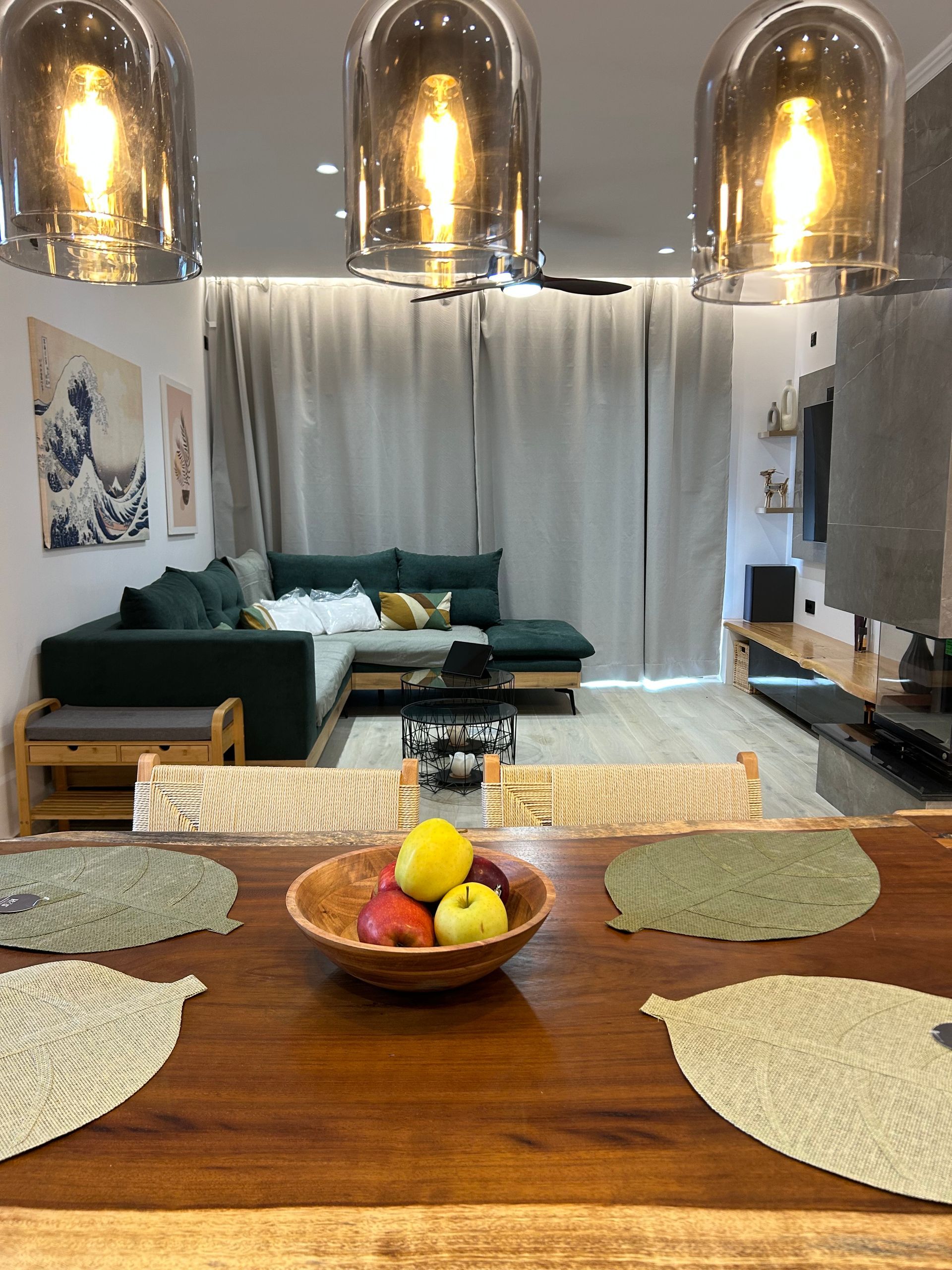 Modern living room with green sectional, pendant lights, curtained window, and a fruit bowl on a wooden table