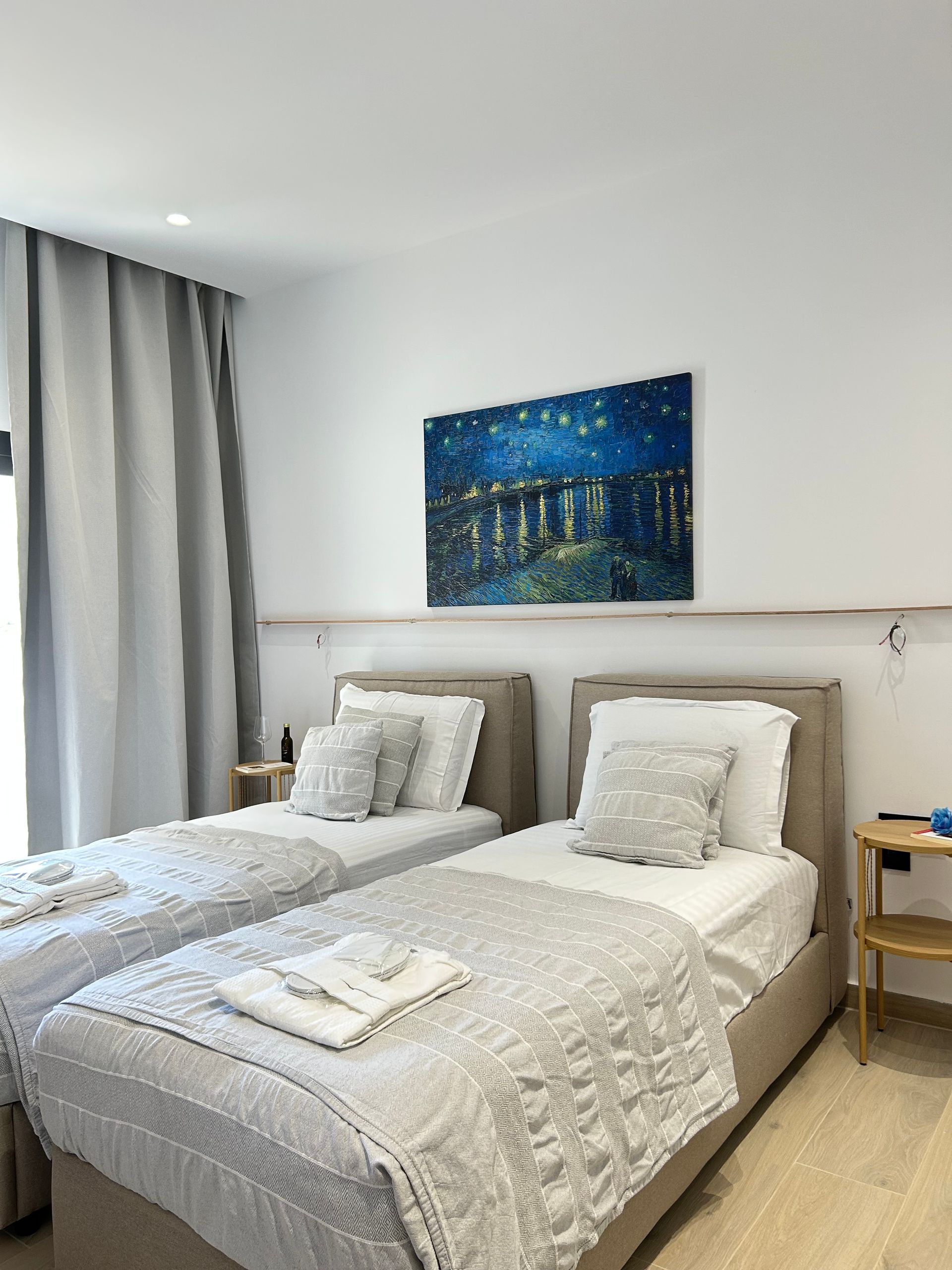 Bright bedroom with two twin beds, white bedding, gray curtains, and a blue painting above the headboard.
