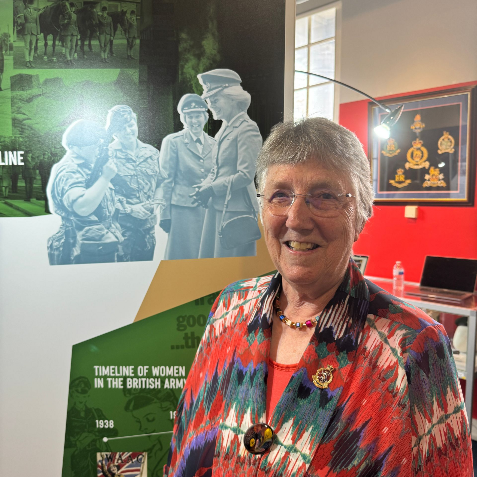 Smiling woman in glasses stands before a museum display about women in the British Army.