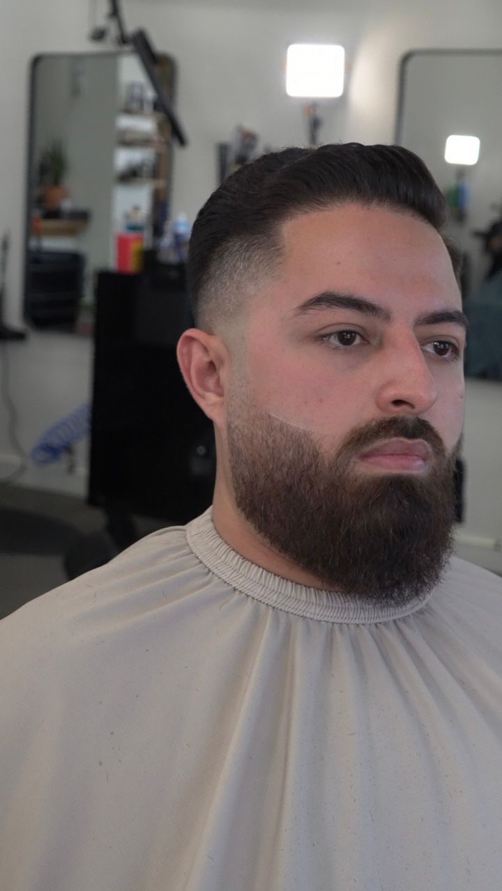 Man with a trimmed beard and fade haircut wearing a cream barber cape in a salon chair