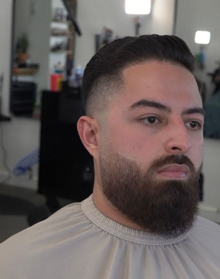 Man with a full beard and slicked-back hair in a barber chair, wearing a beige cape.