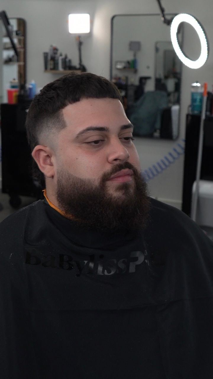 Man with dark beard in a black cape at a barbershop, looking left beside a lit ring light.