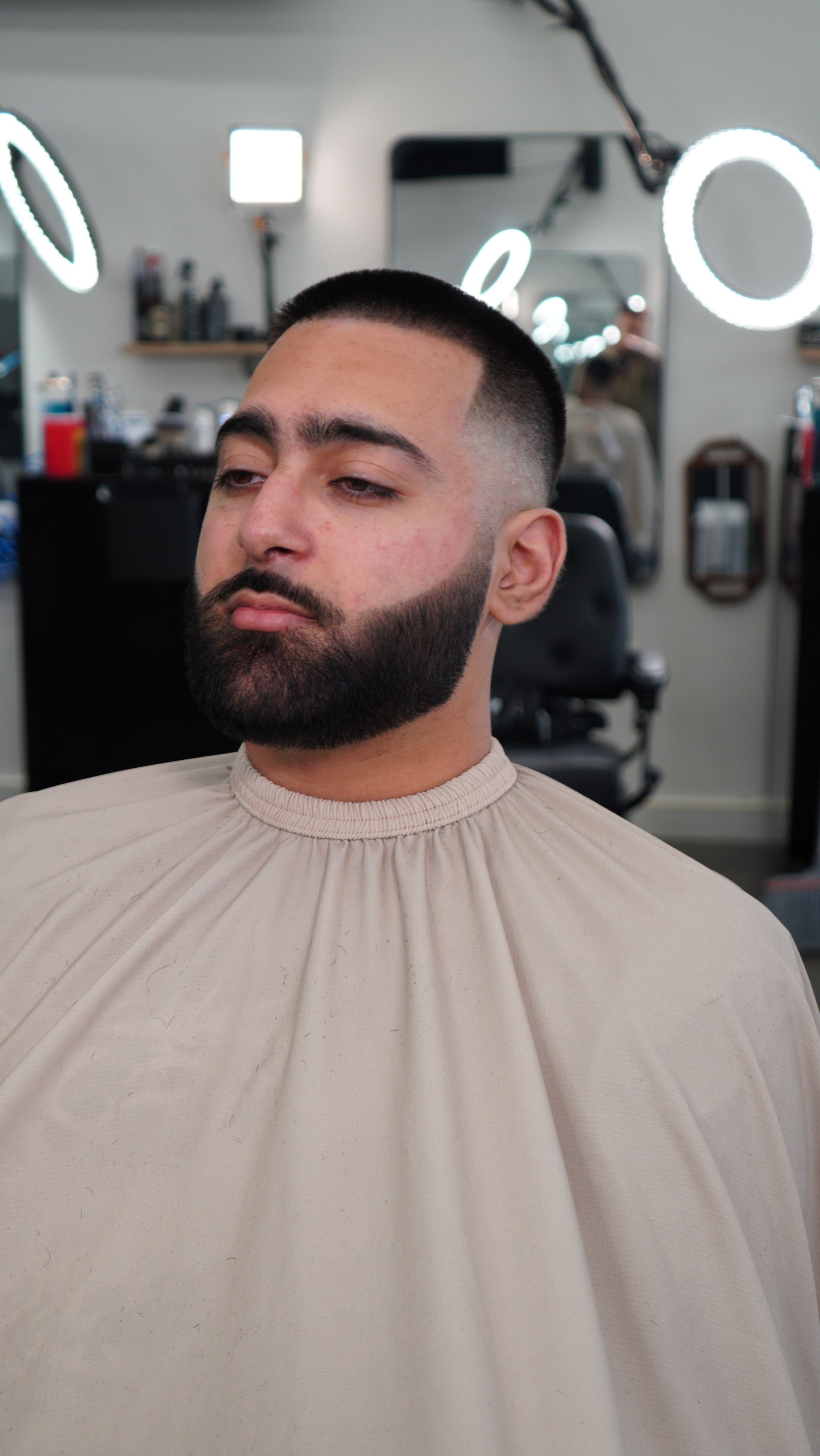 Bearded man in a beige barber cape, seated in a barbershop with ring lights in the background