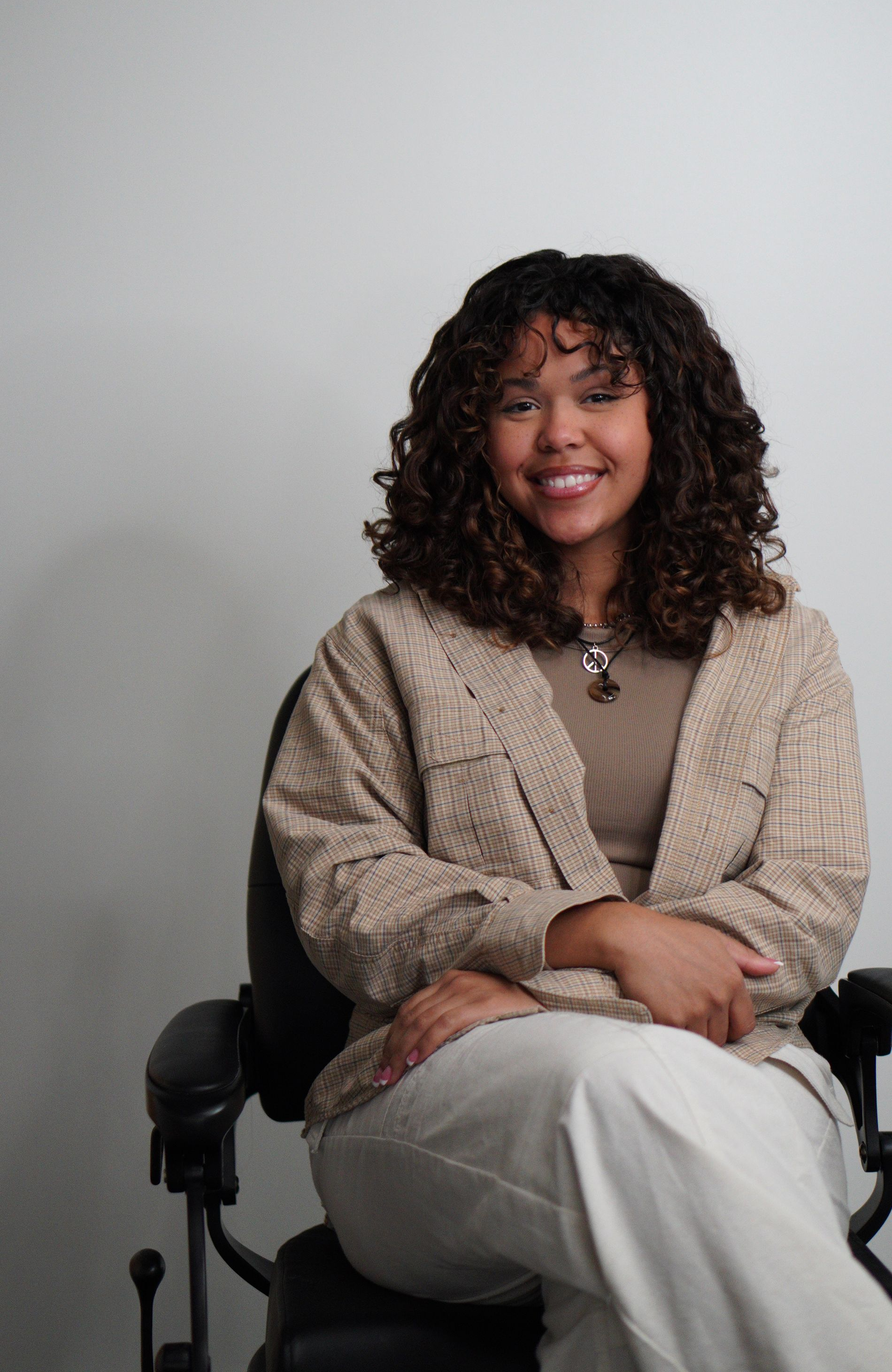 Woman seated in a black chair, smiling in a beige sweater and white pants against a plain gray background