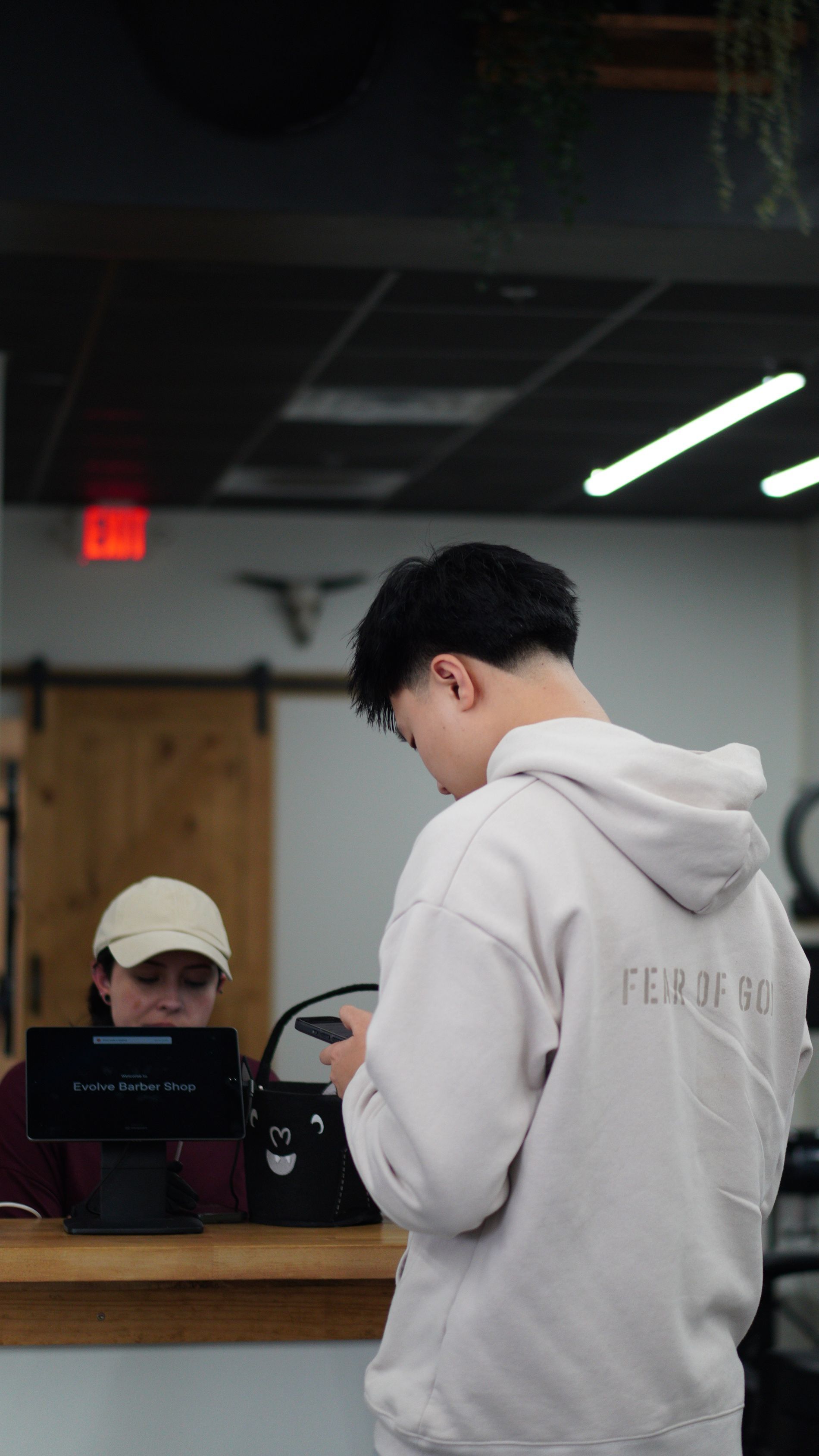 Person in a gray hoodie stands at a counter with a seated clerk in a beige cap inside a dim office.