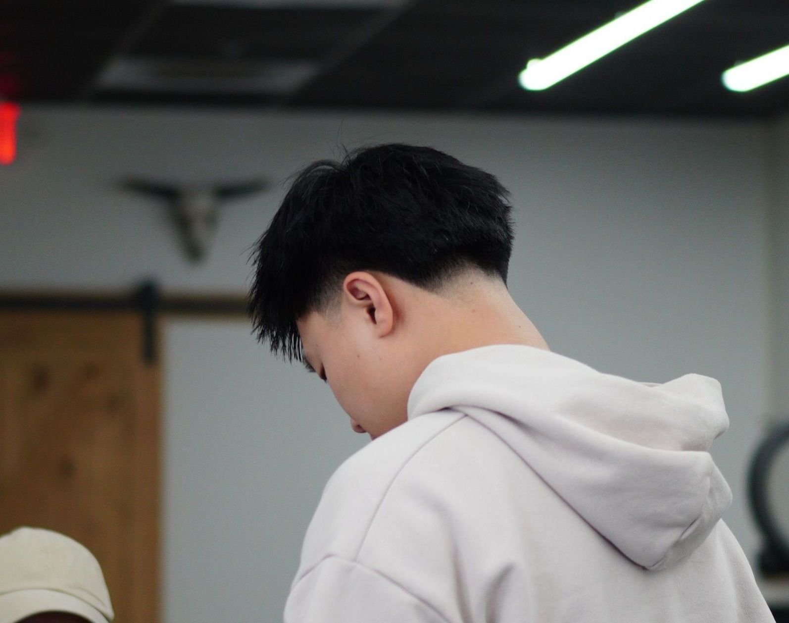Person in a light gray hoodie, viewed from behind, looking down in a softly lit indoor room.