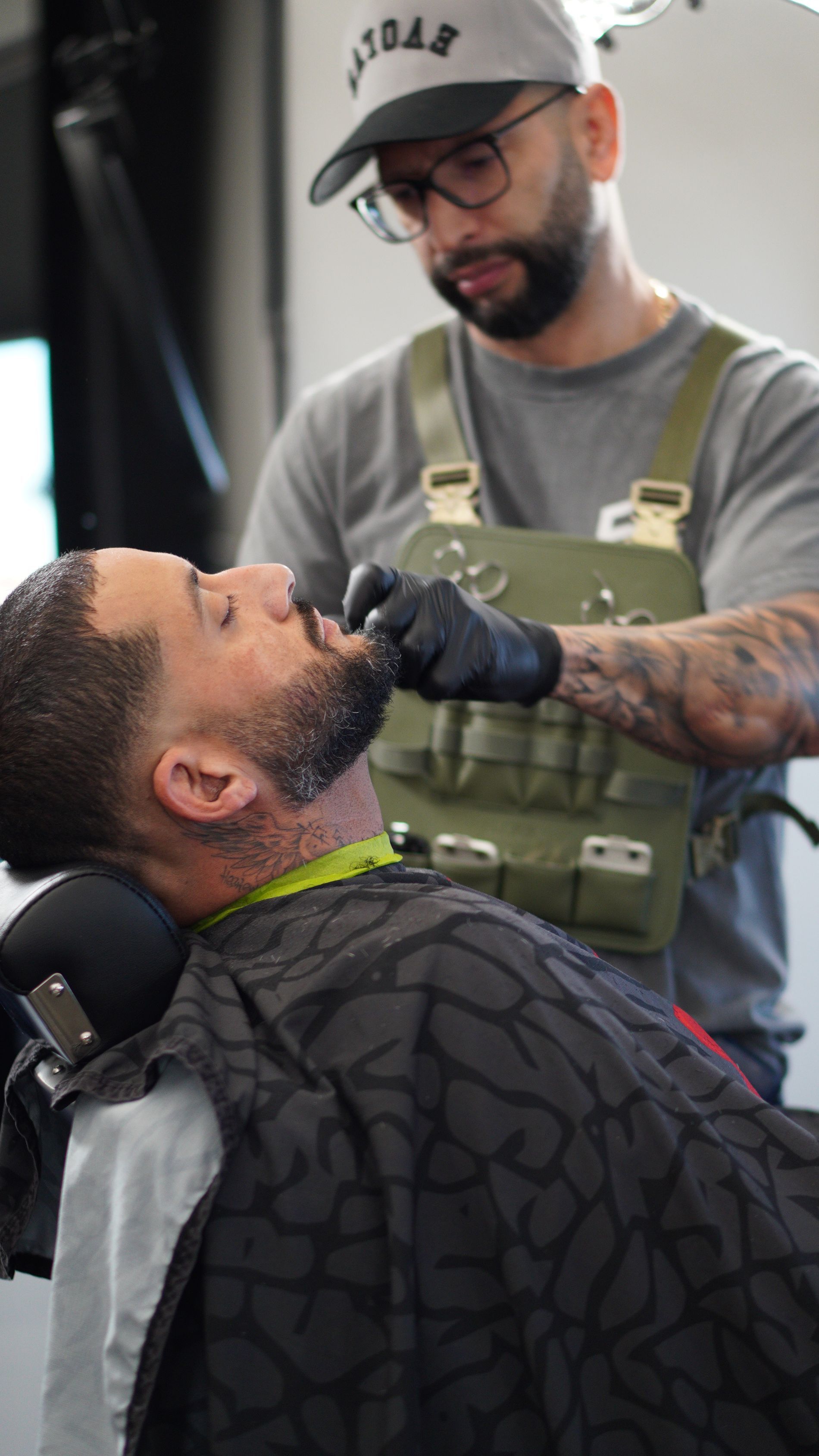 Barber trimming a man's beard with clippers while he wears a cape and neck strip