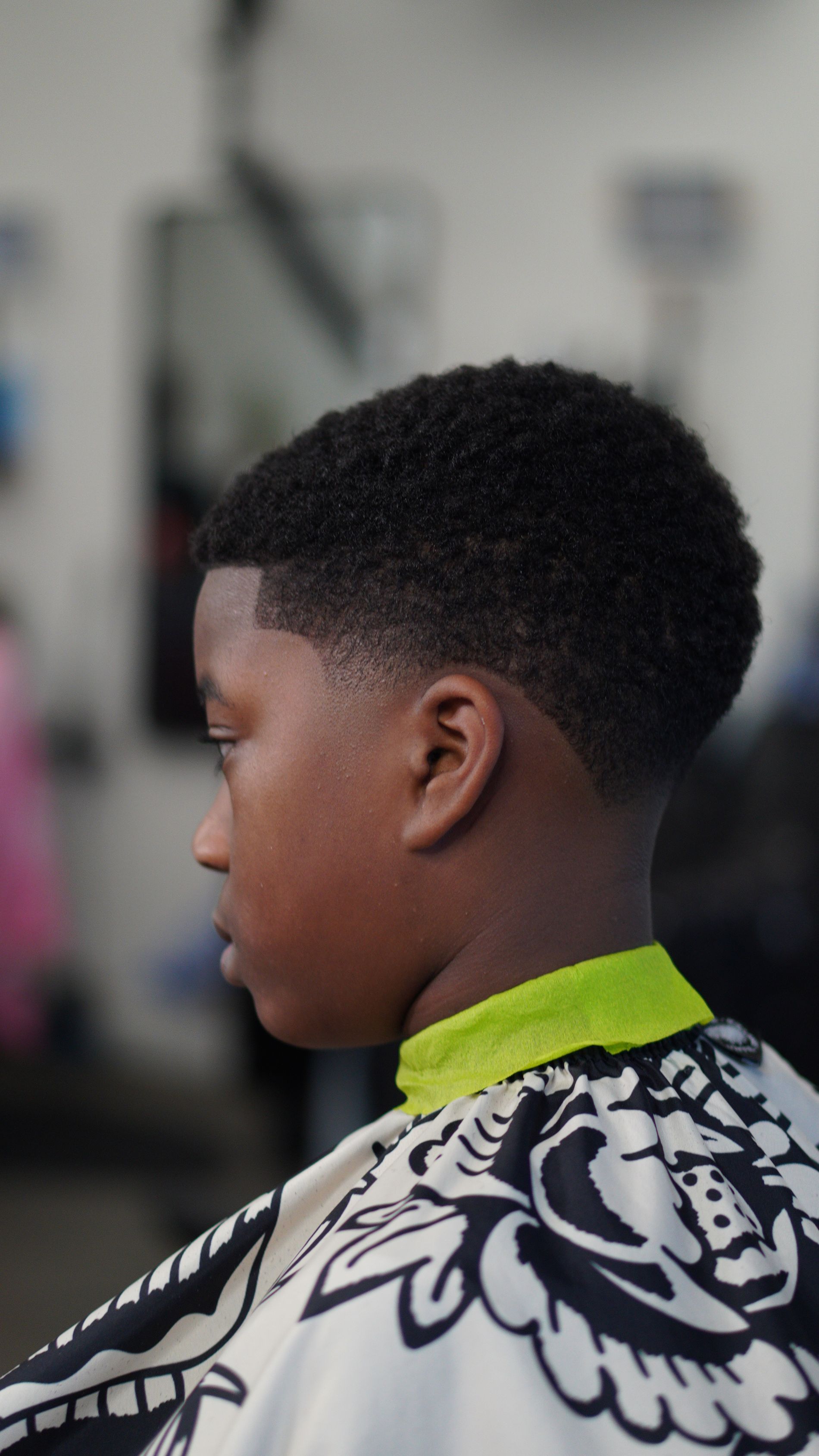 Profile of a person with short curly hair in a patterned cape, seated in a salon chair.
