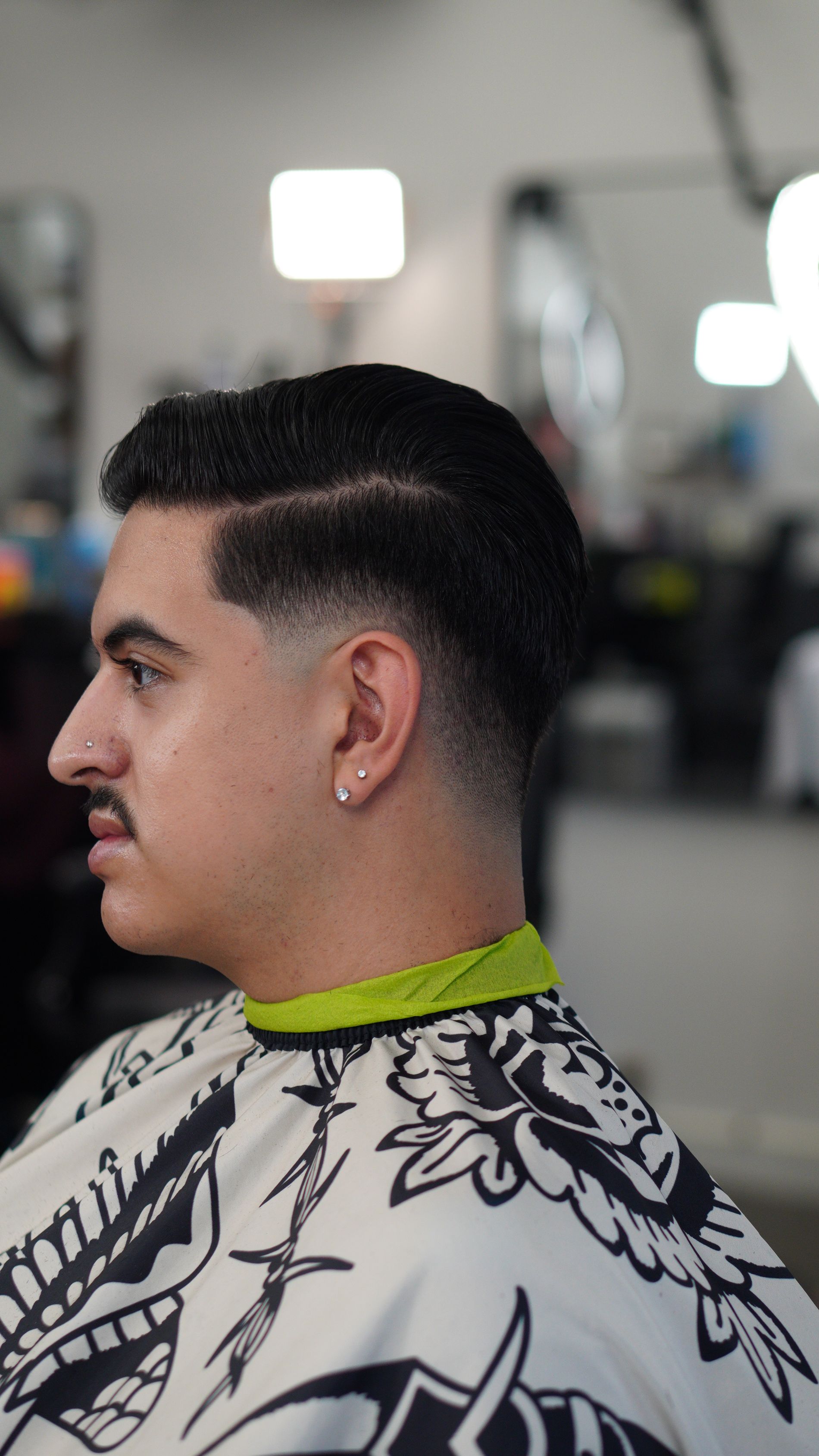 Side profile of a man in a barber chair wearing a black-and-white cape in a barbershop.