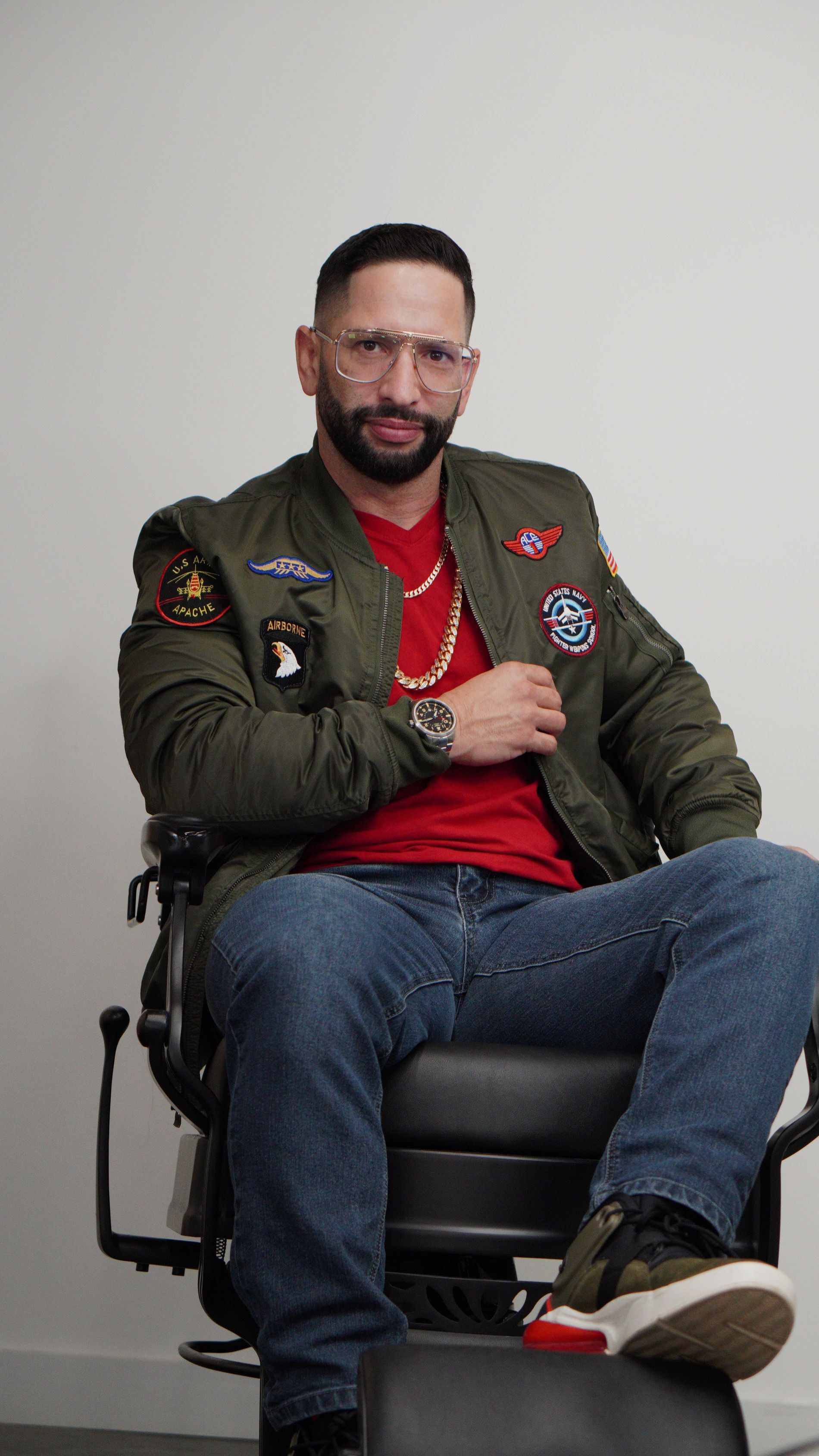 Man seated on a chair, wearing an olive jacket, red shirt, and jeans, against a plain gray background