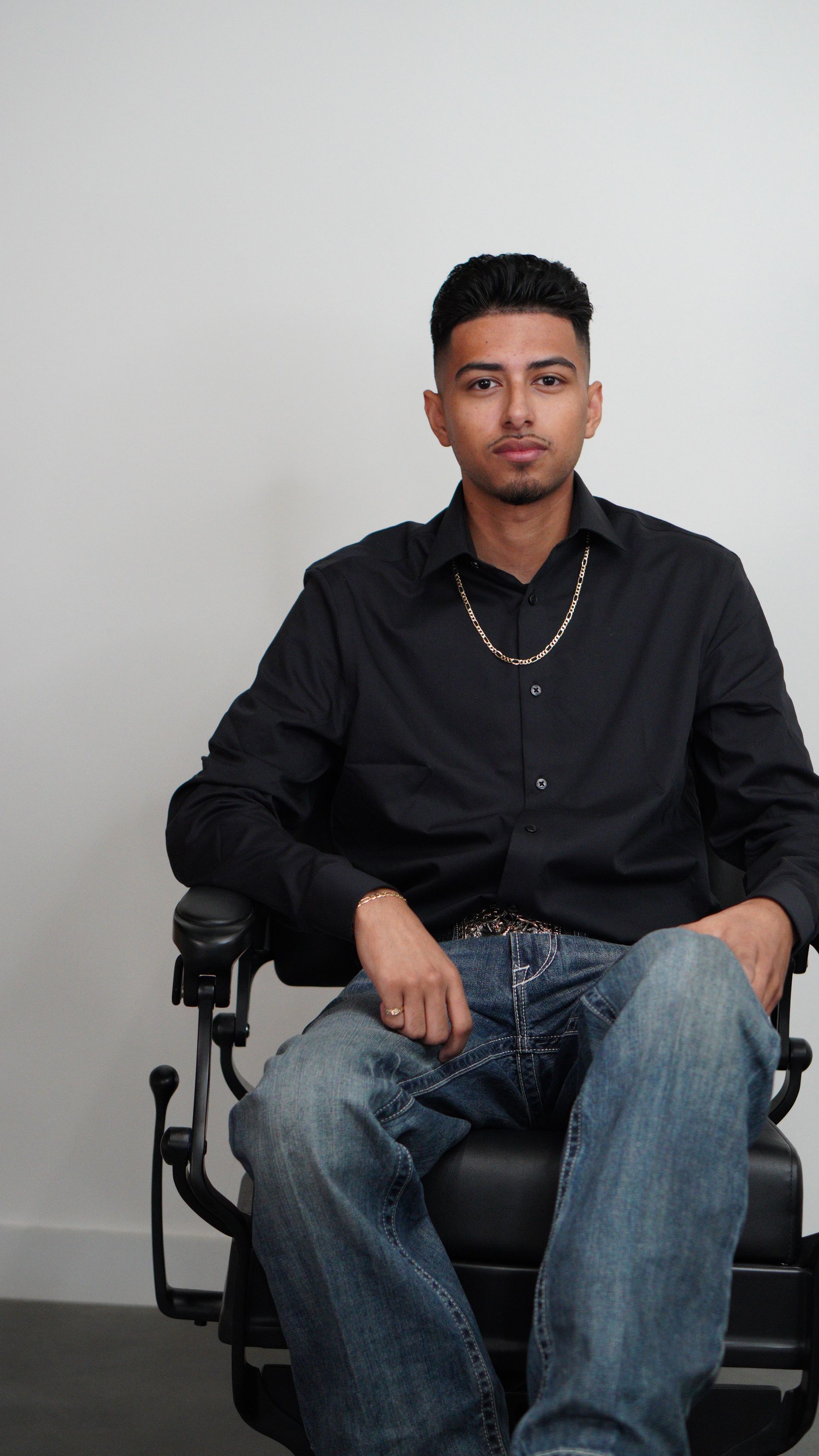Man seated on a black office chair against a plain light wall, wearing a black shirt and jeans.