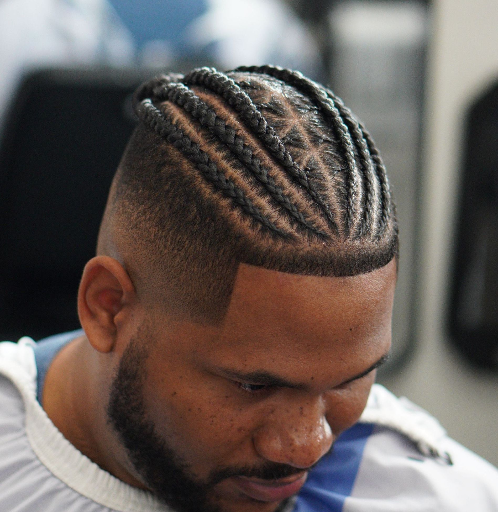Man with braided cornrows looking down in a barber chair, wearing a white cape.