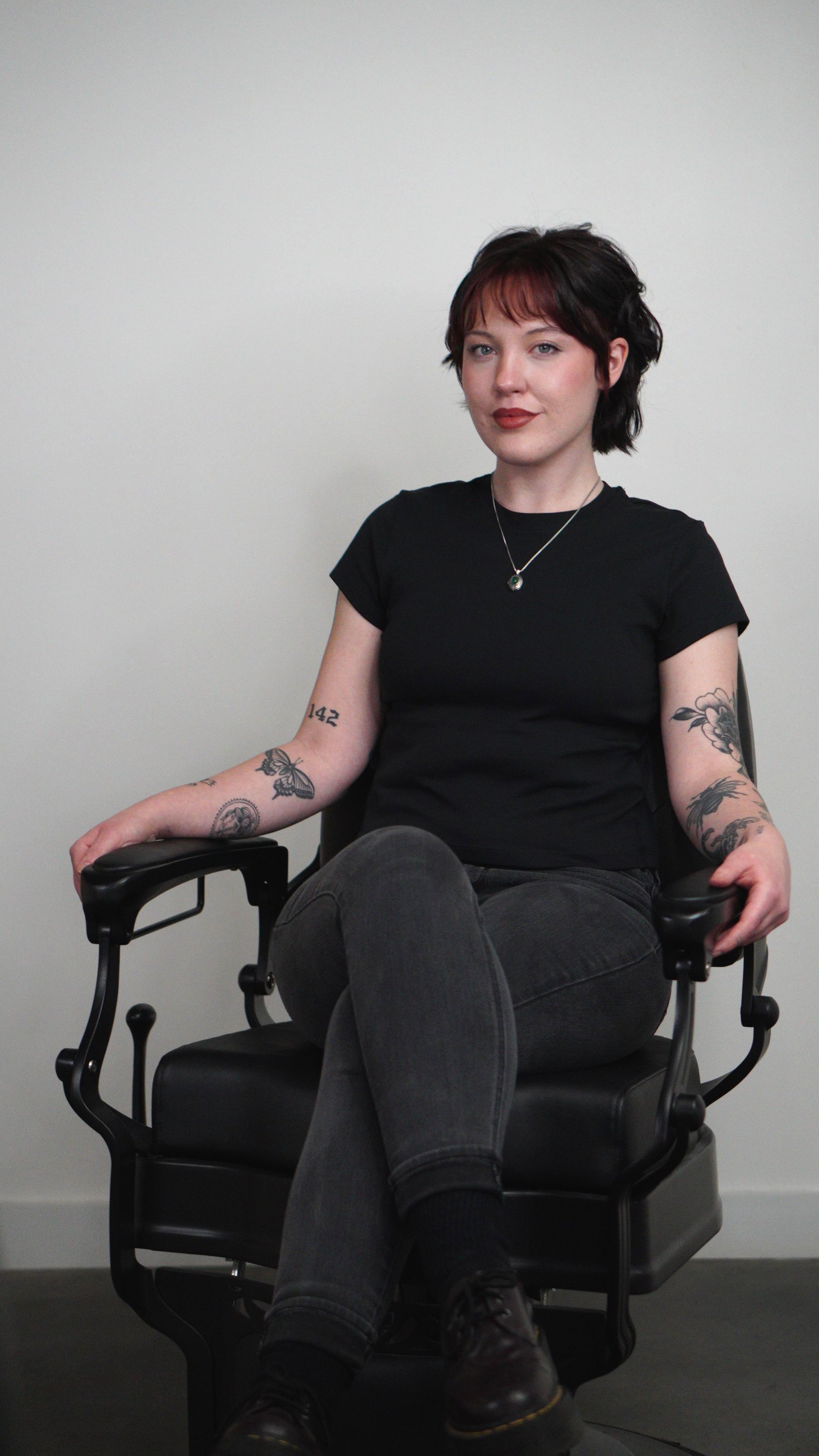 Person seated in a black chair against a plain wall, wearing a black shirt and dark jeans, with visible arm tattoos