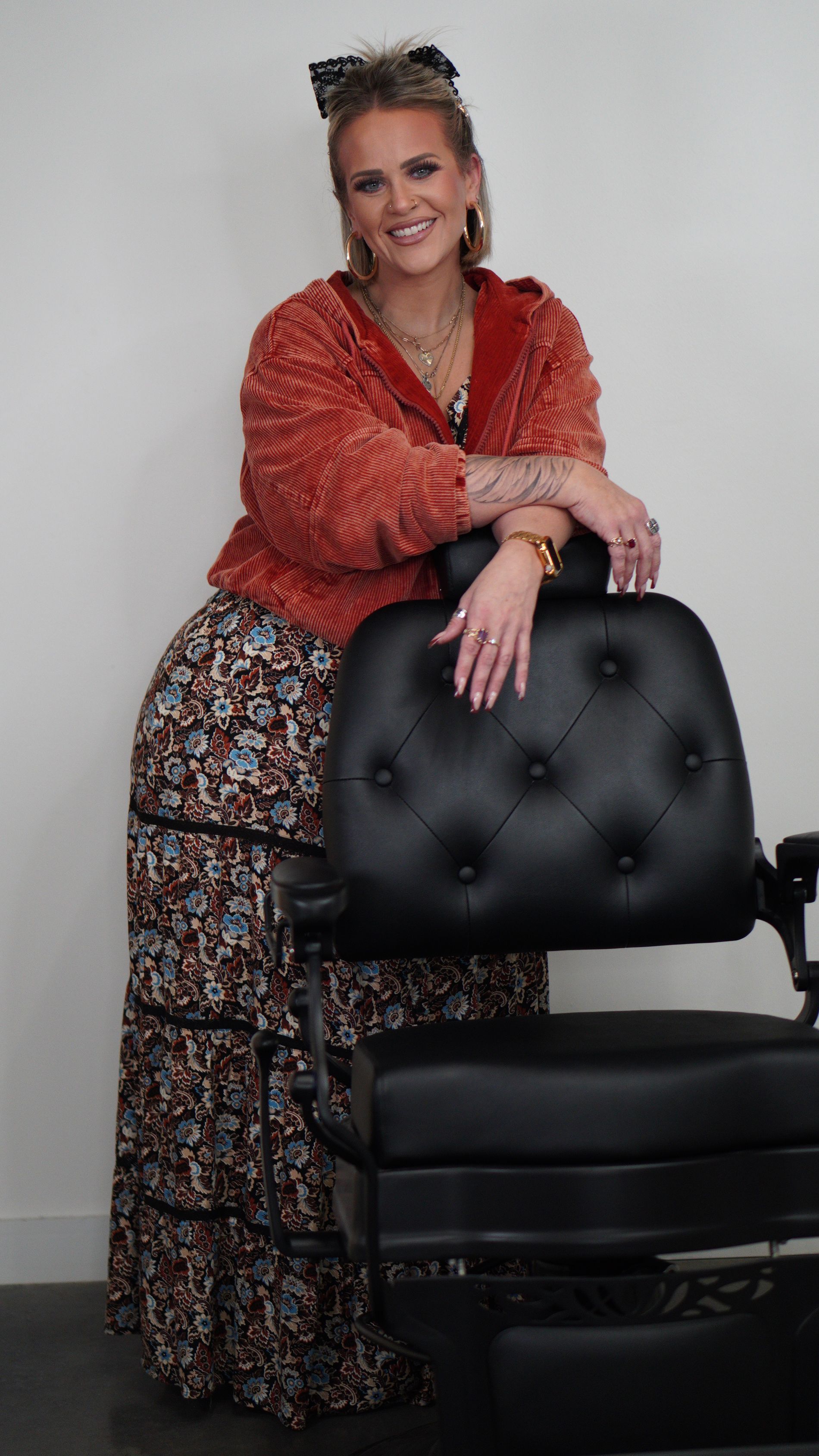 Woman in a red blouse and patterned pants leaning on a black office chair, smiling against a gray backdrop