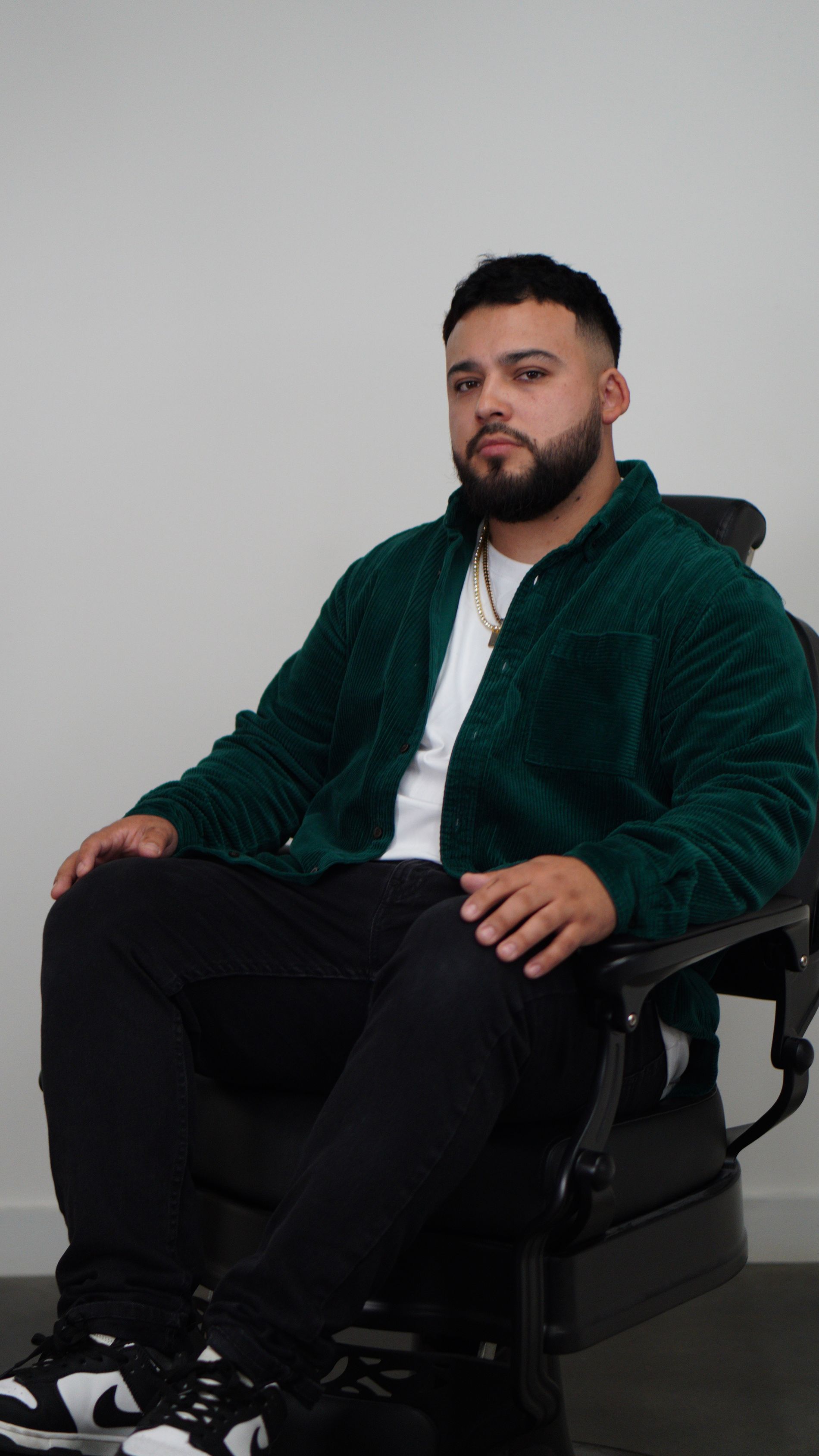 Man seated in a chair wearing a dark green jacket, white shirt, and black pants against a plain wall.
