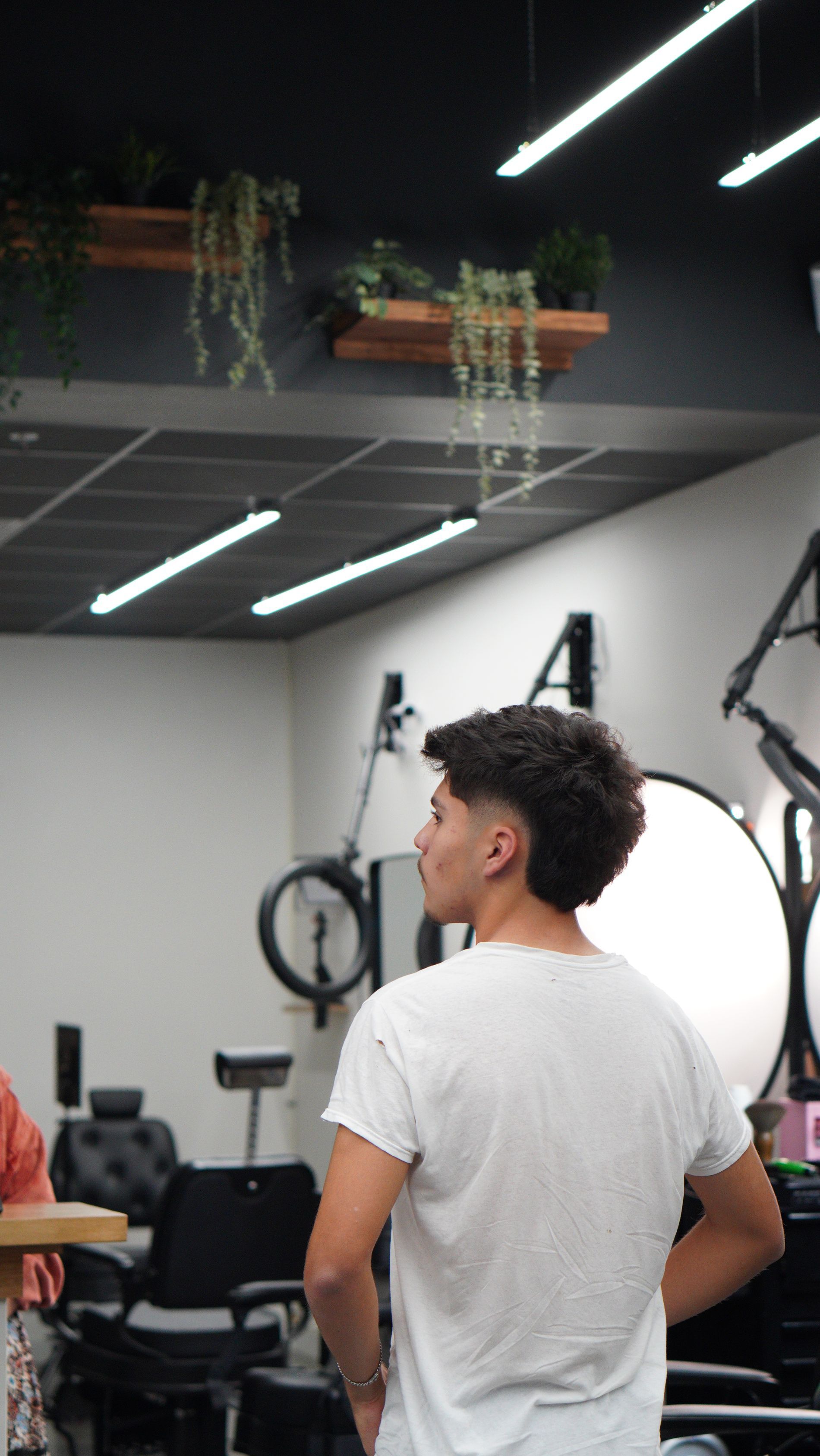 Person in a white T-shirt stands in a gym/barber studio, seen from behind near mirrors and lighting.