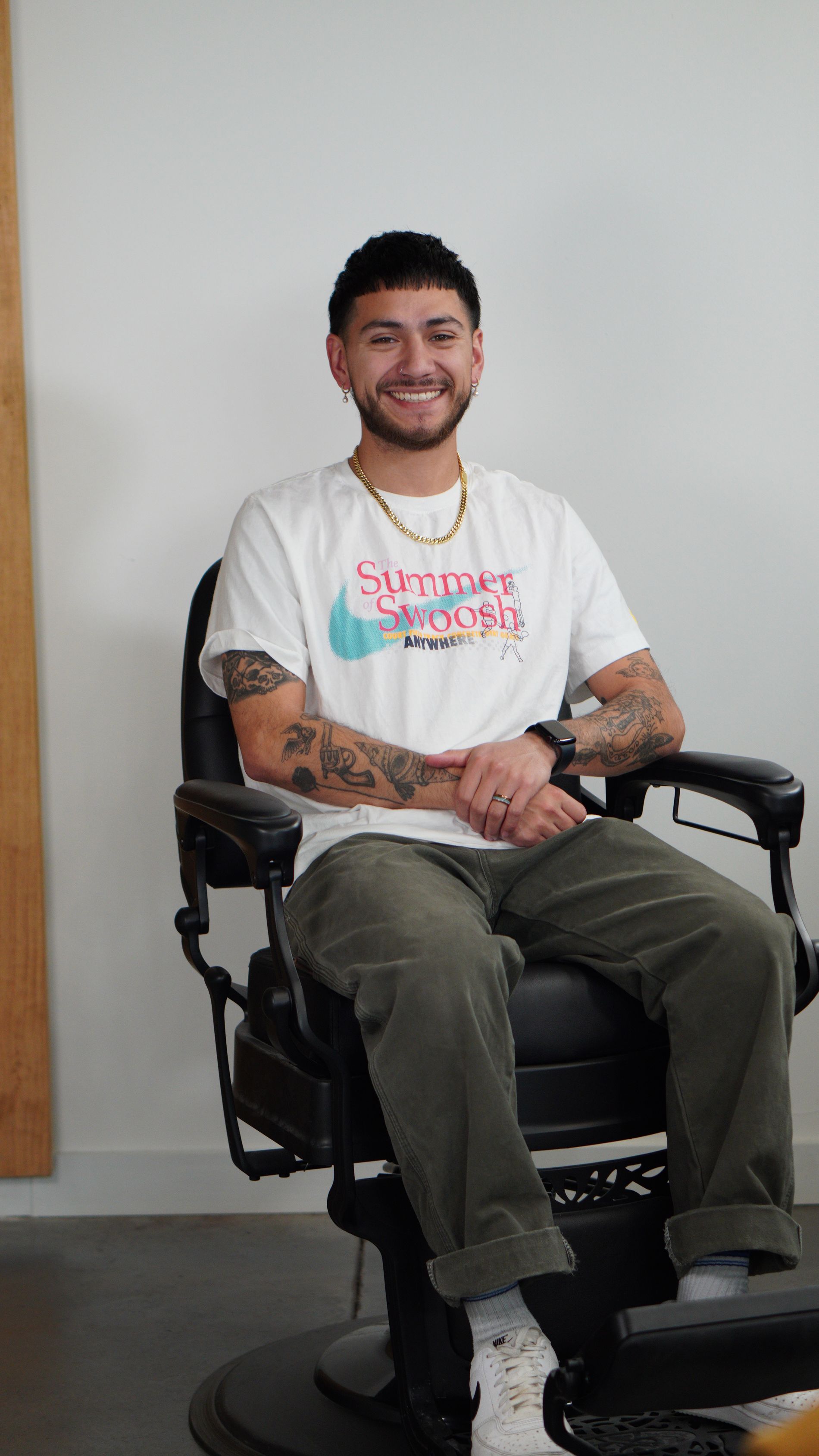 Person smiling in a wheelchair, wearing a white graphic T-shirt and olive pants, indoors against a plain wall.