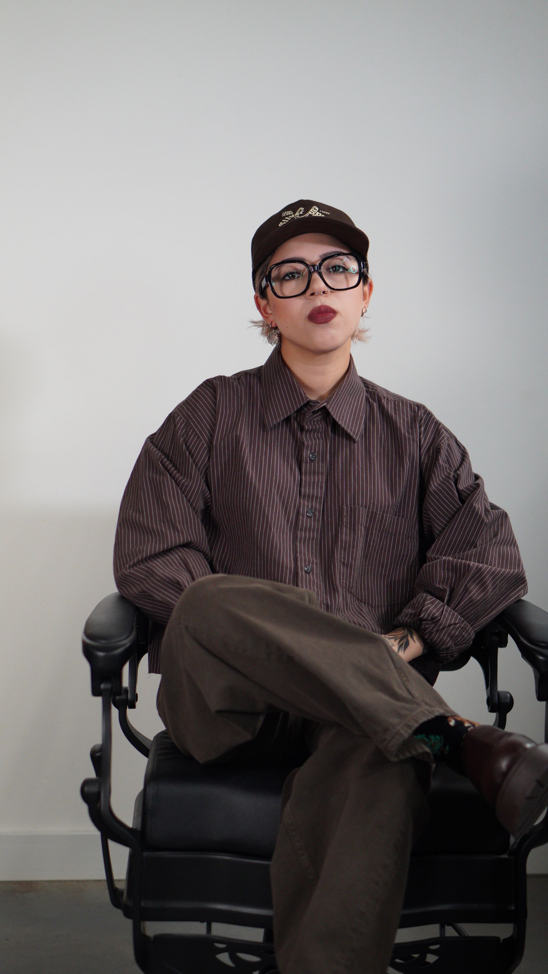 Person seated in a black office chair, wearing a patterned shirt, brown pants, cap, and glasses, against a plain wall.