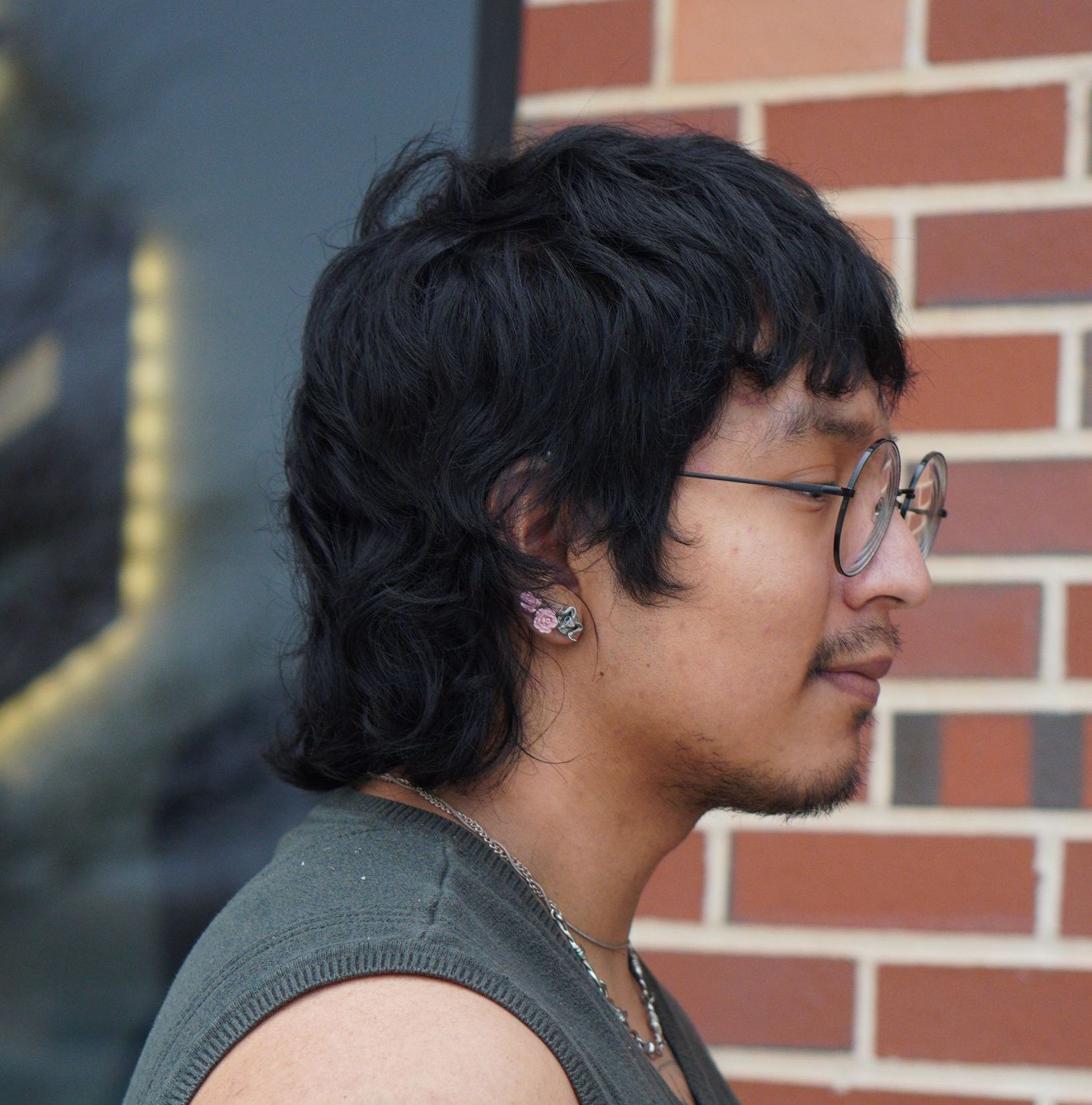 Profile view of a person with glasses and dark hair standing by a brick wall outdoors