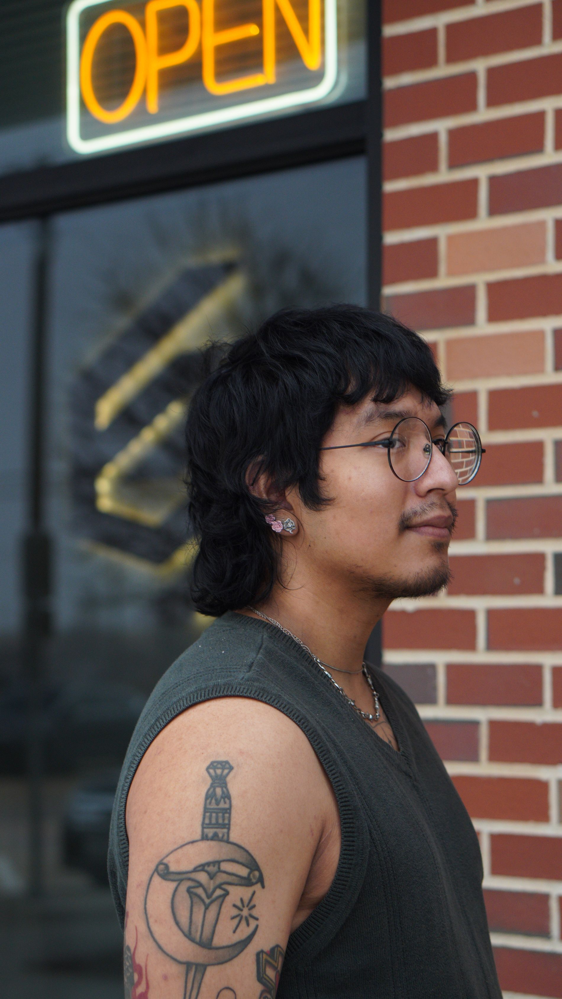 Person with glasses and arm tattoo standing by a brick wall under a neon 