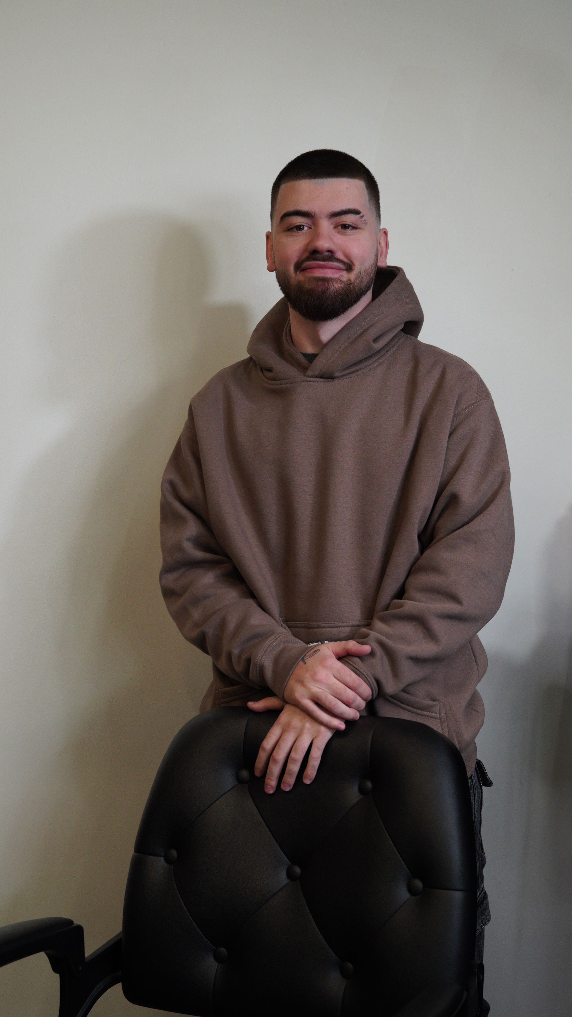 Man in a brown hoodie seated on a black chair against a plain wall, looking at the camera.