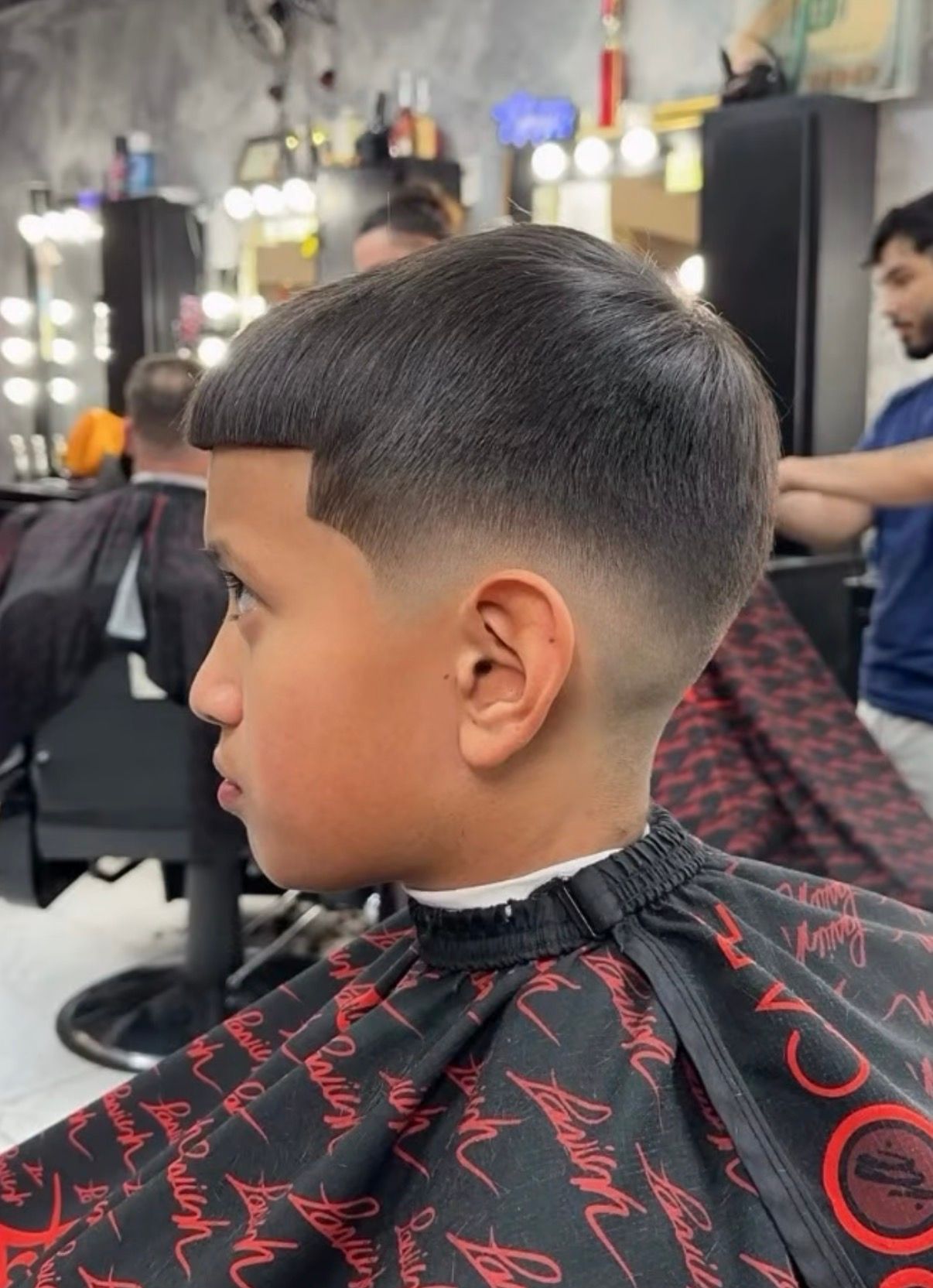 Boy with a fresh fade haircut sitting in a barber chair, wearing a black cape with red patterns.
