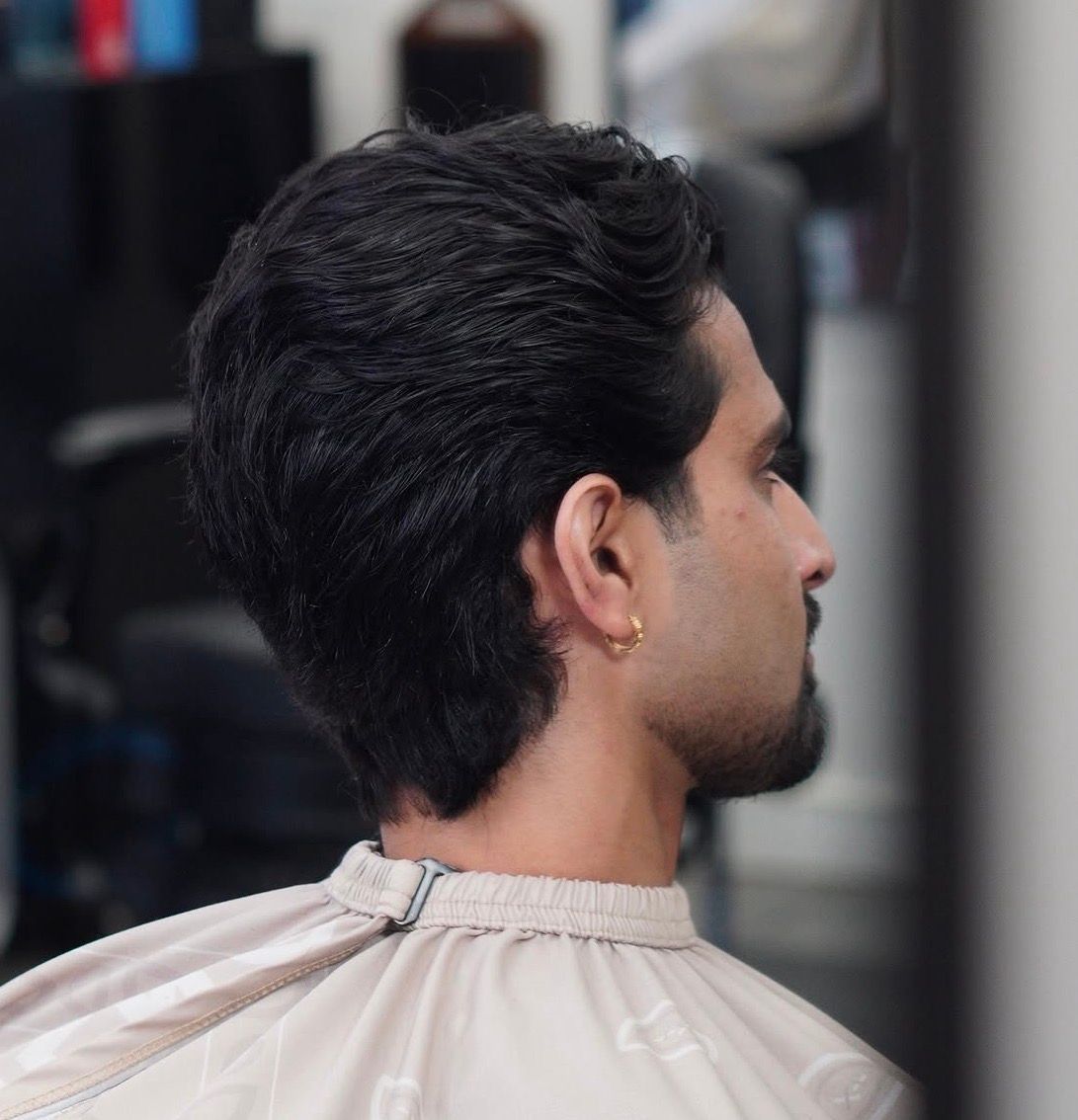 Side profile of a man with dark hair at a barber chair, wearing a light cape, in a salon.