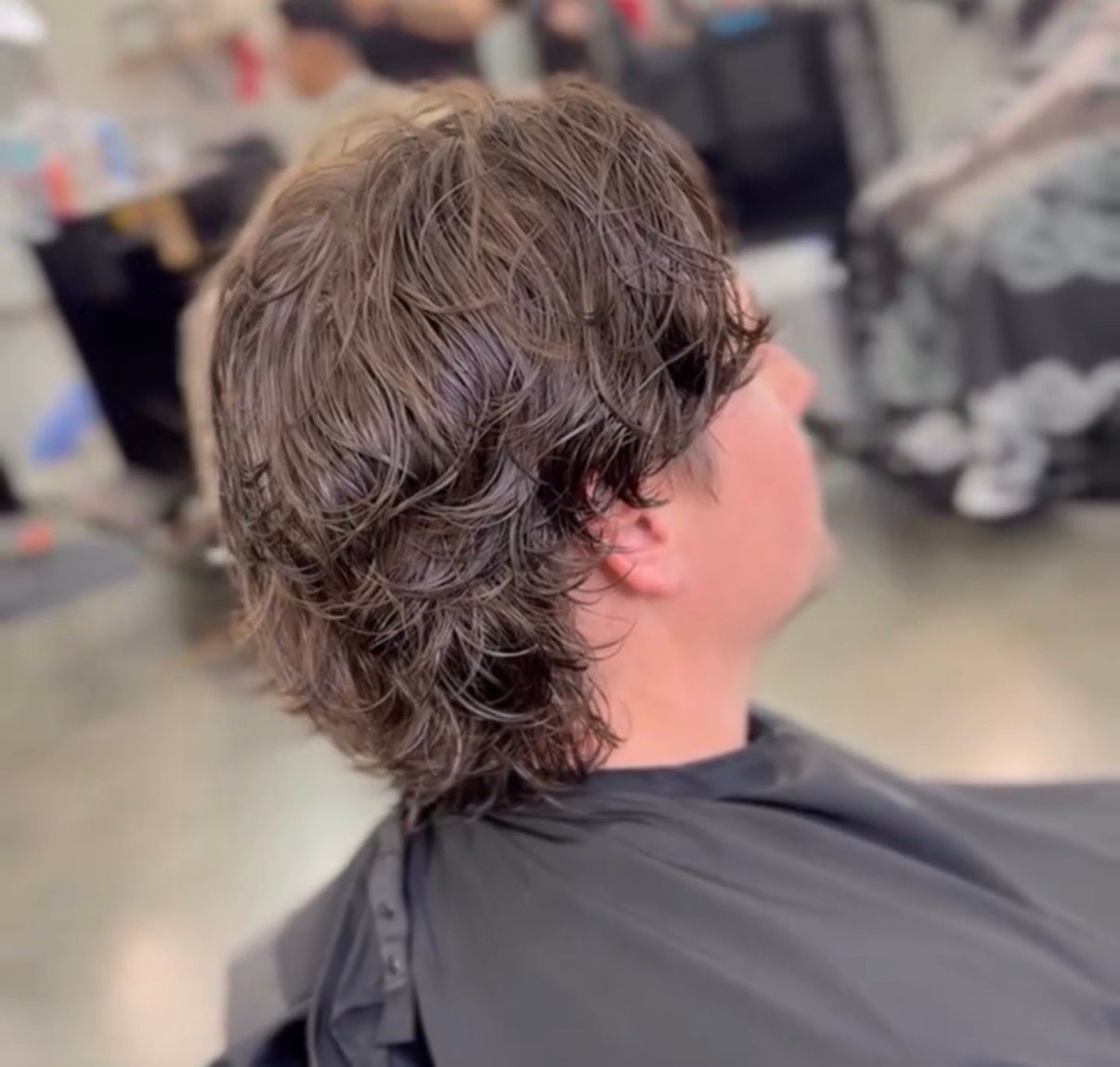 Person with wavy brown hair sitting in a salon chair, viewed from the side.