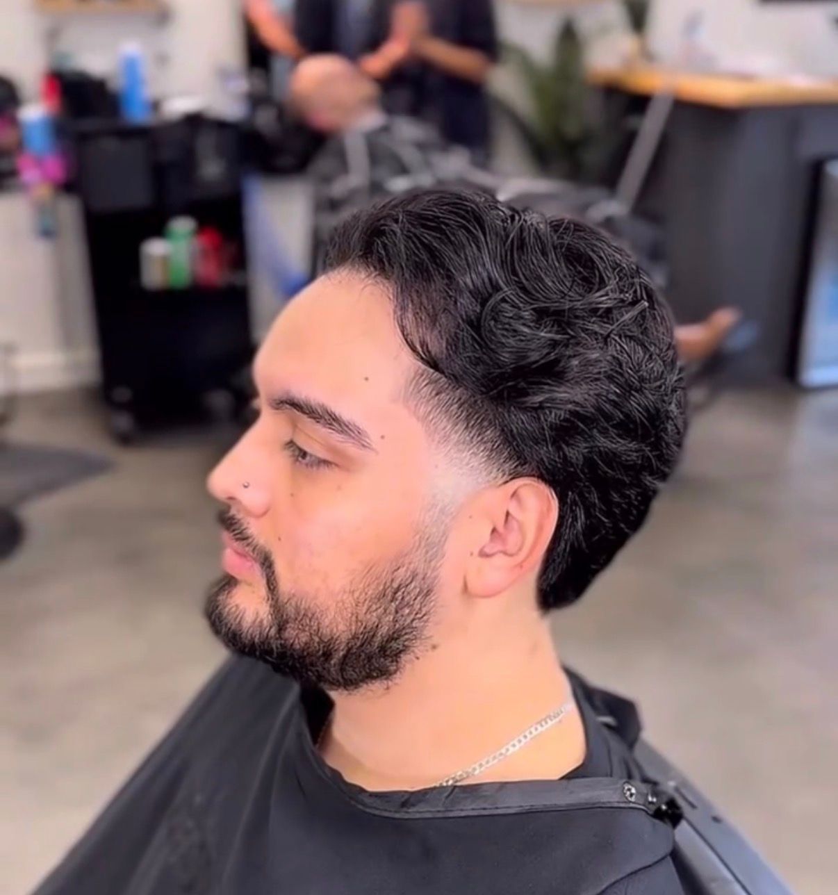 Side-profile of a man with a trimmed beard and dark curly hair in a barbershop chair, showing a fresh fade haircut
